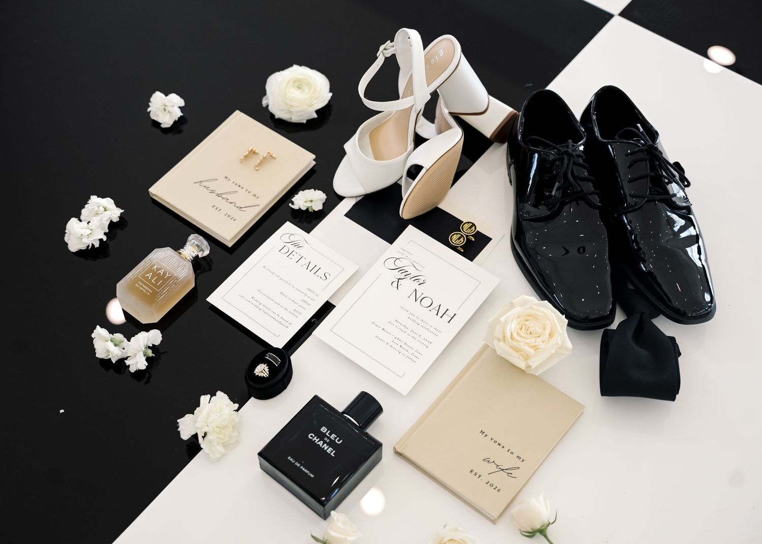 Elegant wedding flat lay with white heels, black dress shoes, invitations, perfume, and floral accents on checkered floor.