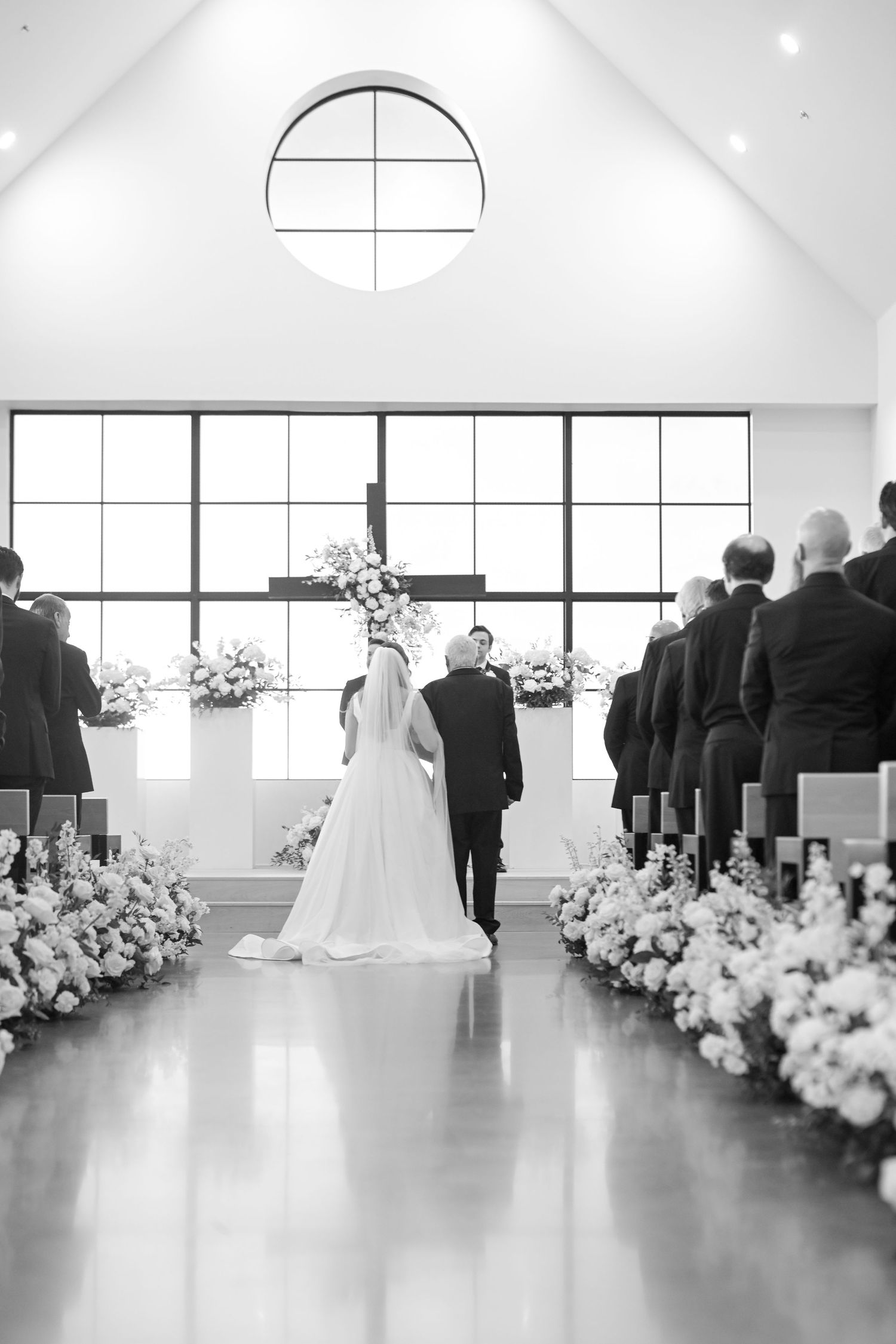 Black and white photo of bride and groom walking down a floral-lined aisle in a modern chapel.