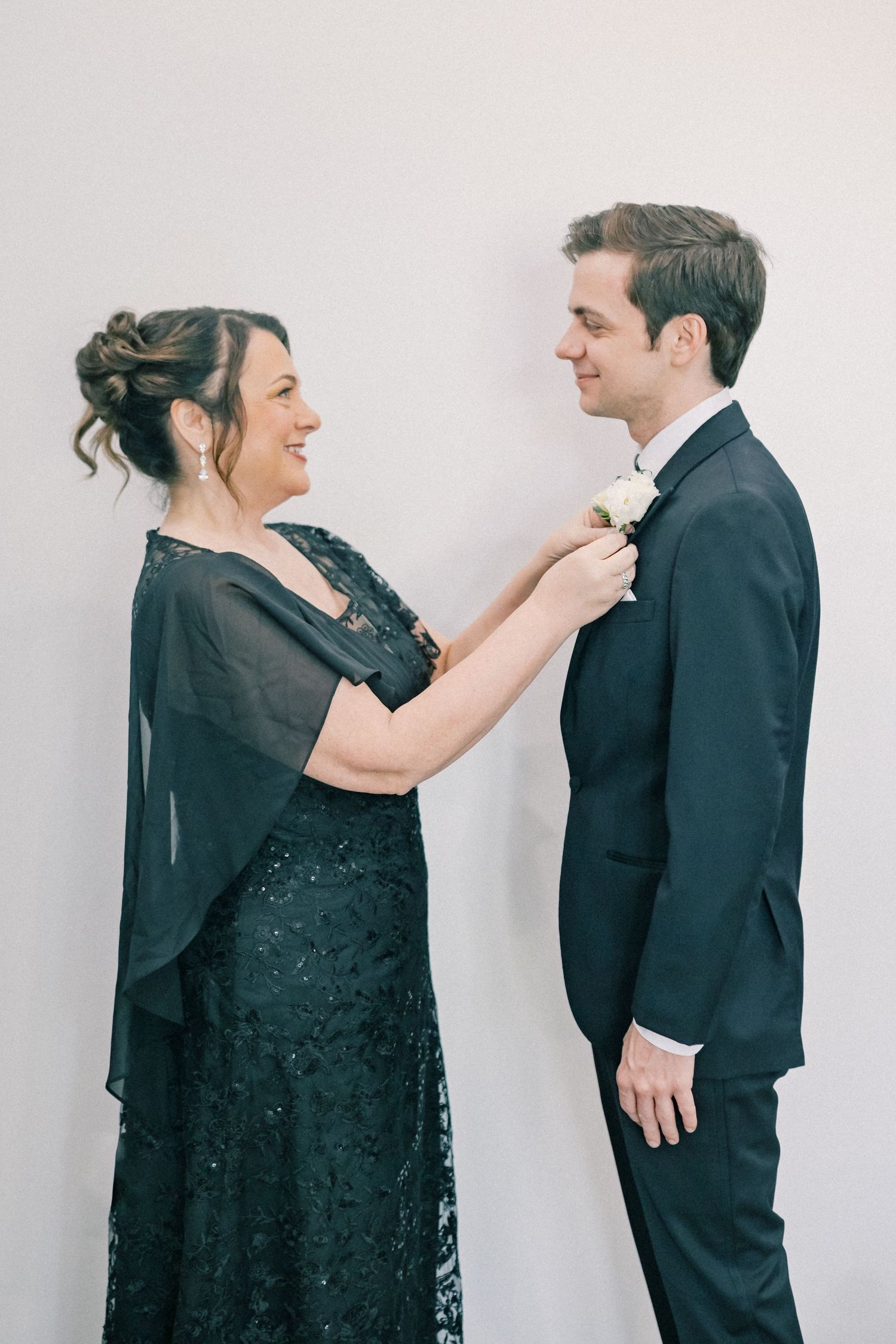Mother of groom pinning white boutonniere on groom in navy suit before wedding.