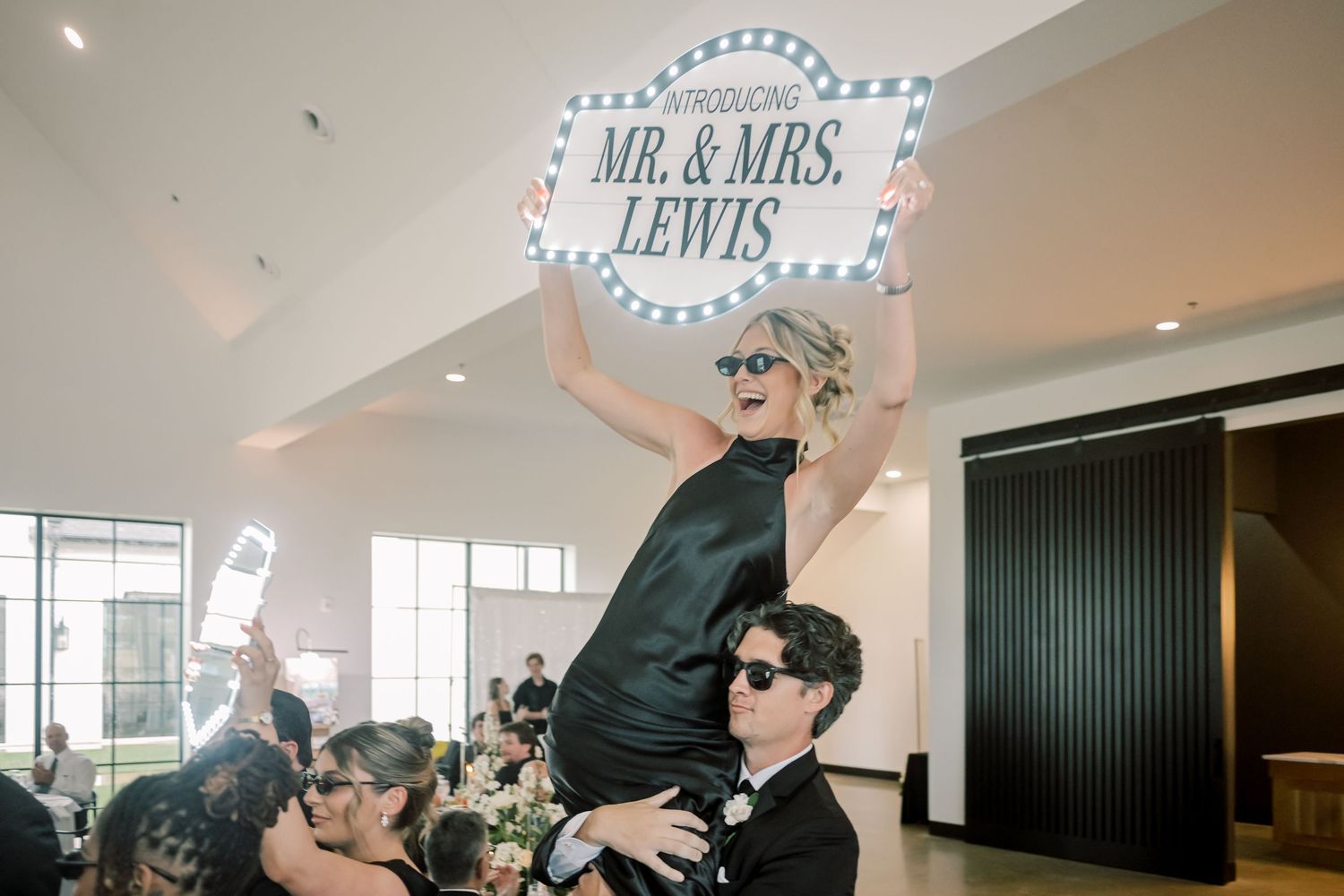 Woman in black dress held up holding a Mr. & Mrs. Lewis sign at a lively wedding reception.