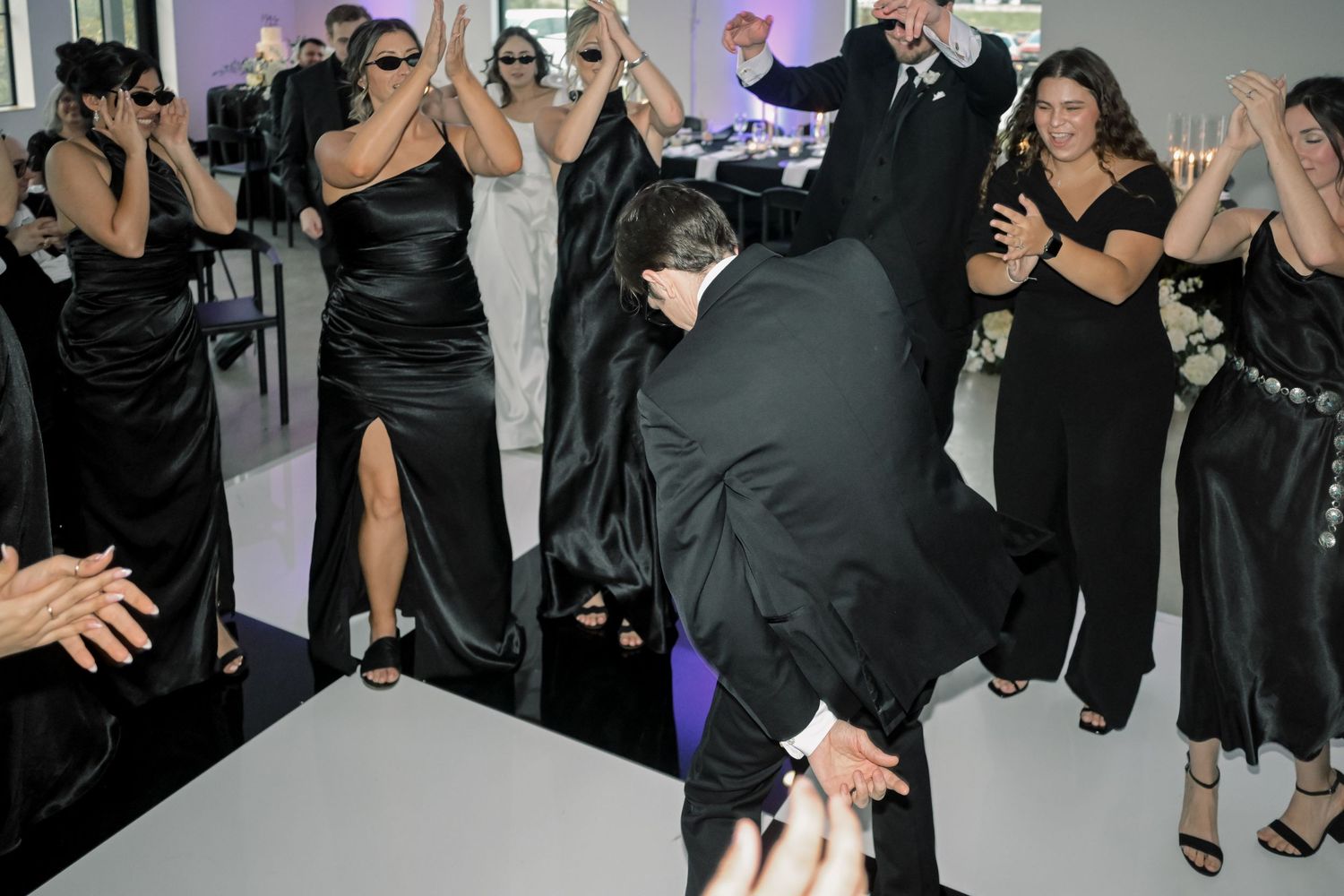 Wedding guests in black formal attire dancing and celebrating energetically on a white dance floor.