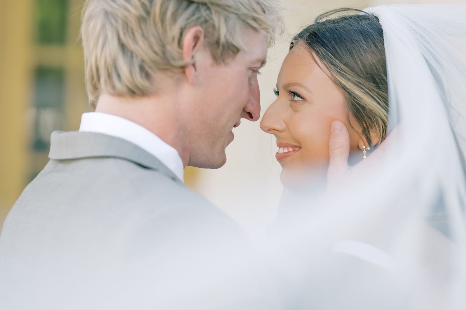 A bride in a white veil and groom in gray suit share an intimate moment, smiling at each other on their wedding day.