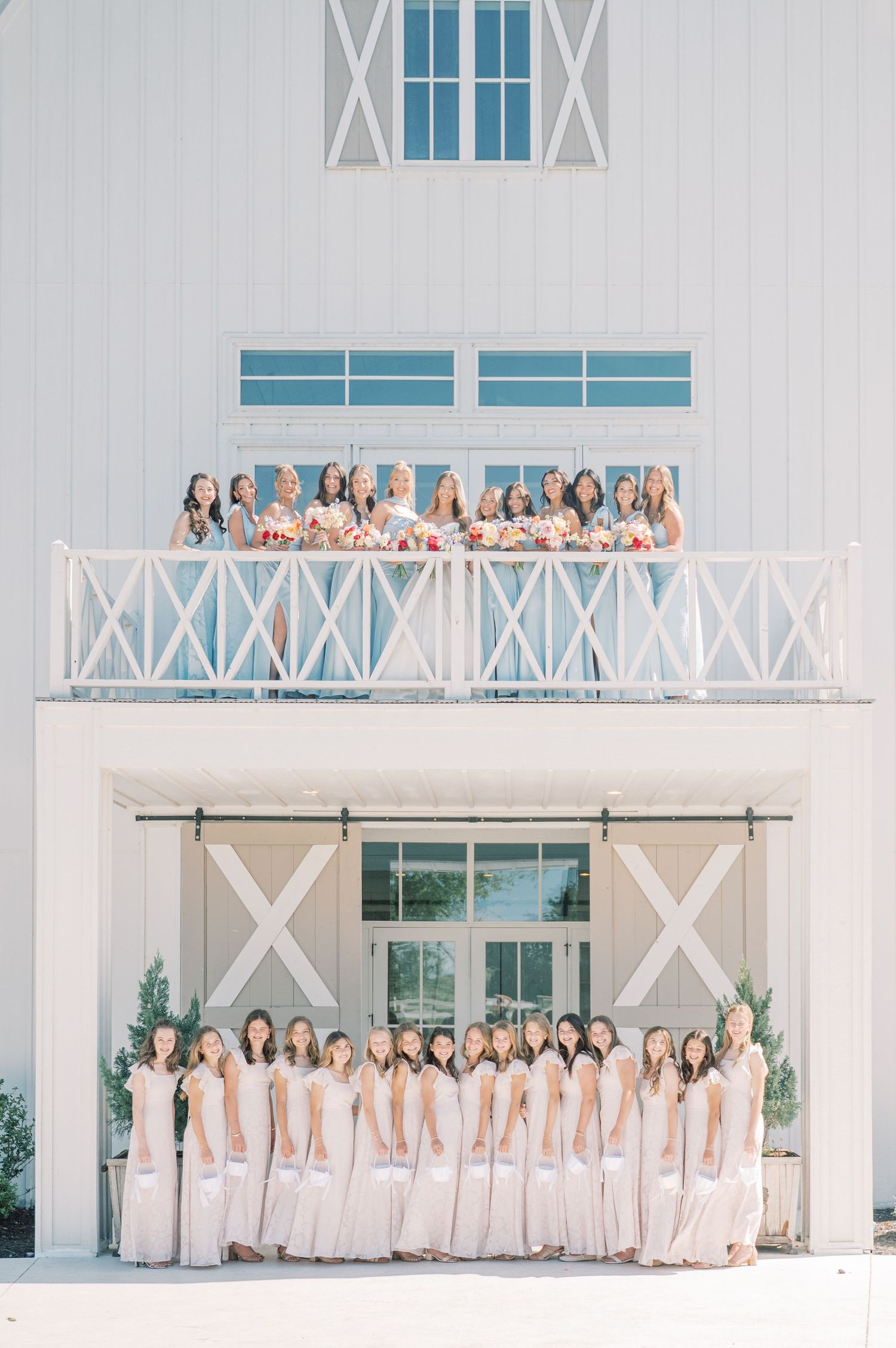 Large bridal party poses in white dresses outside a white barn venue, with bridesmaids on balcony holding bouquets.