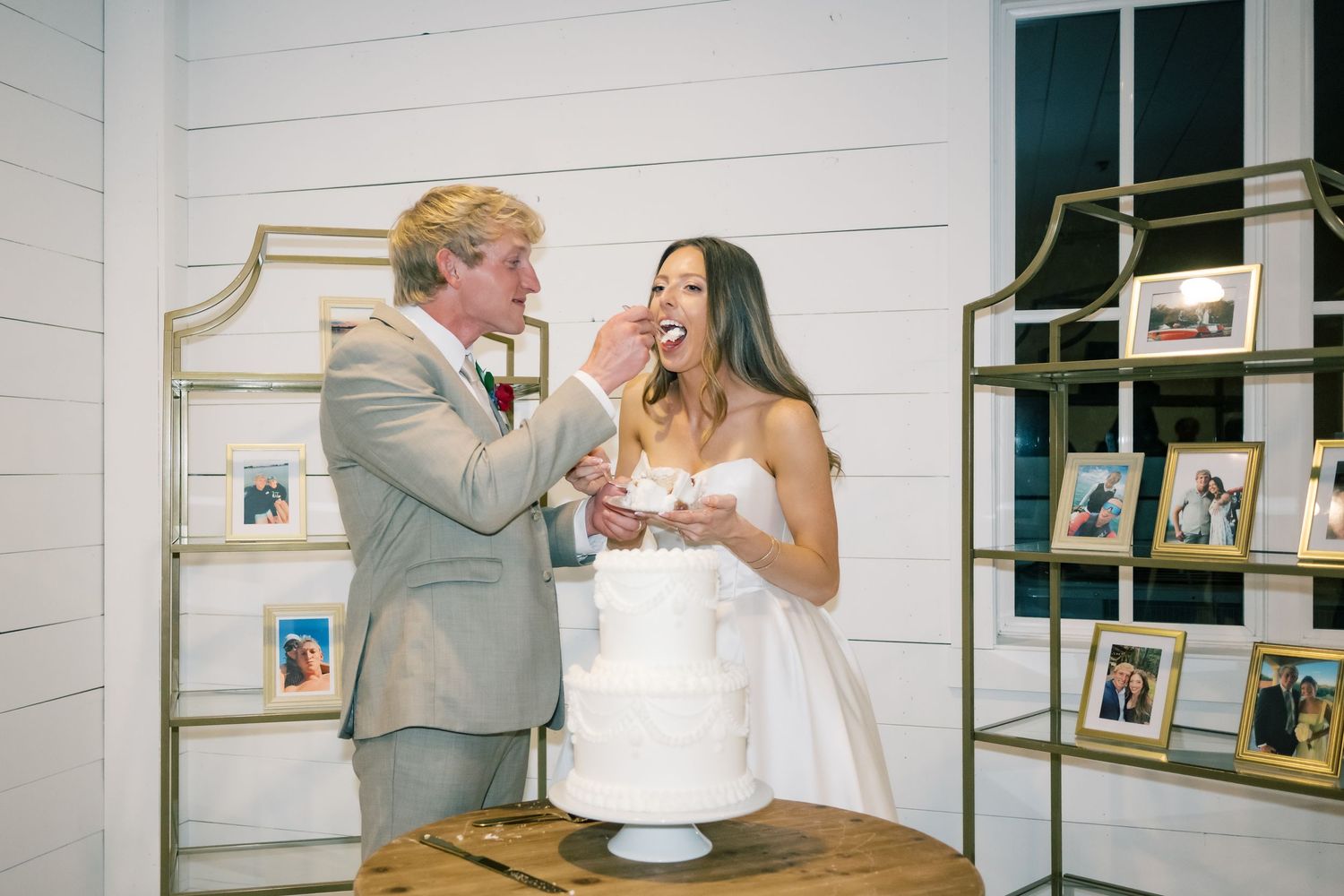 Bride and groom laughing while cutting white wedding cake at rustic venue with gold shelving.