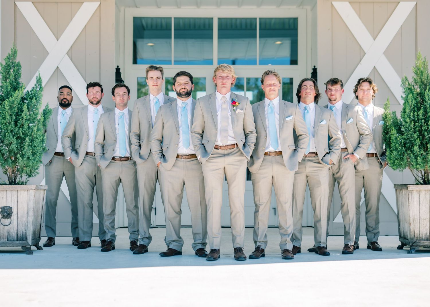 Groomsmen in tan suits with light blue ties pose together outside a white barn venue for a wedding photo.