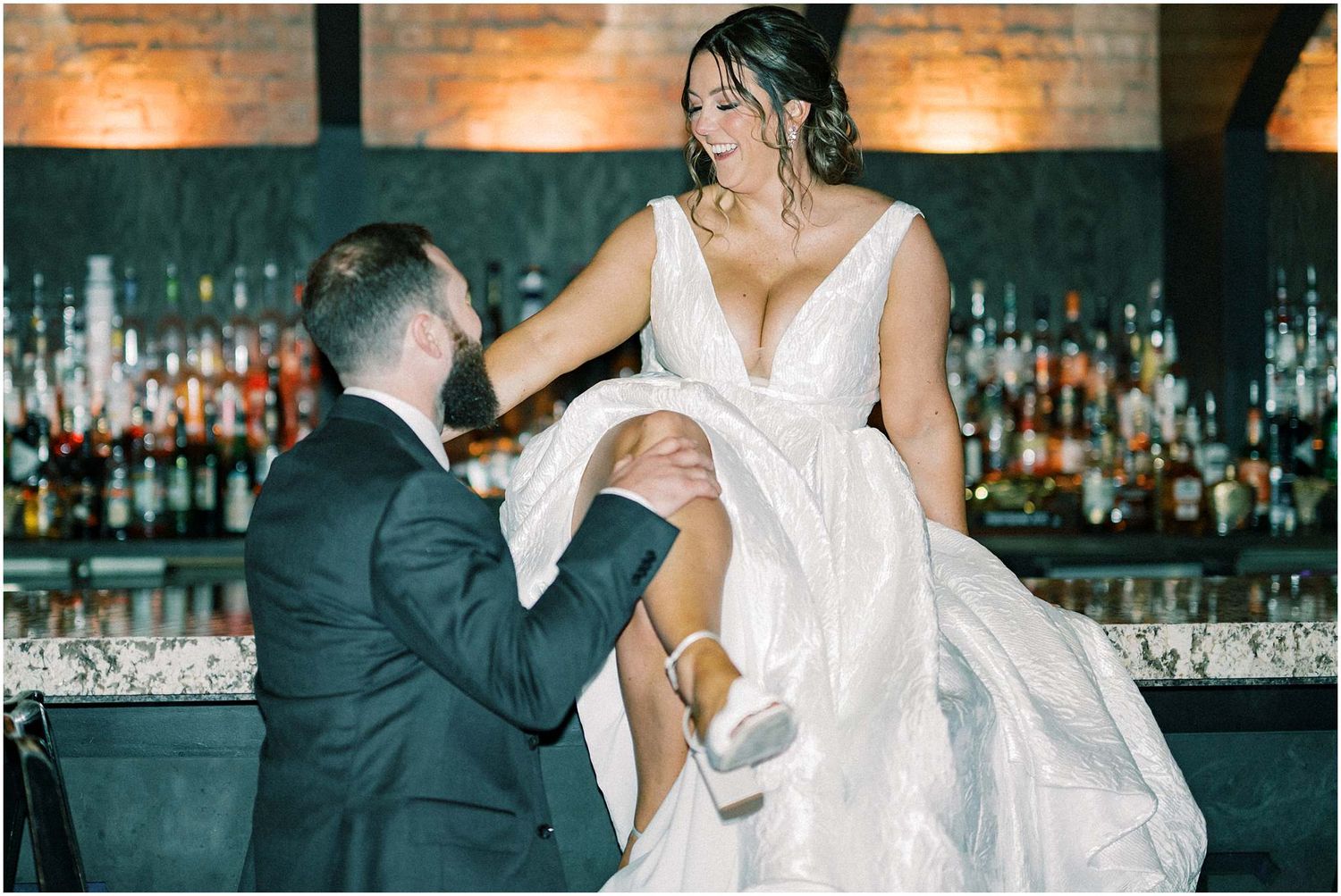 Wedding couple dips and dances together in front of a rustic brick wall with ambient lighting.