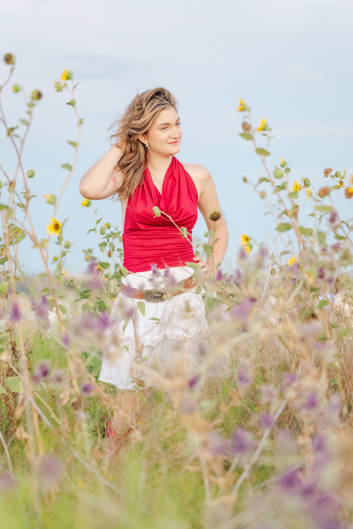 Woman in red halter top and white skirt standing in a wildflower field holding a sunflower on a cloudy day.