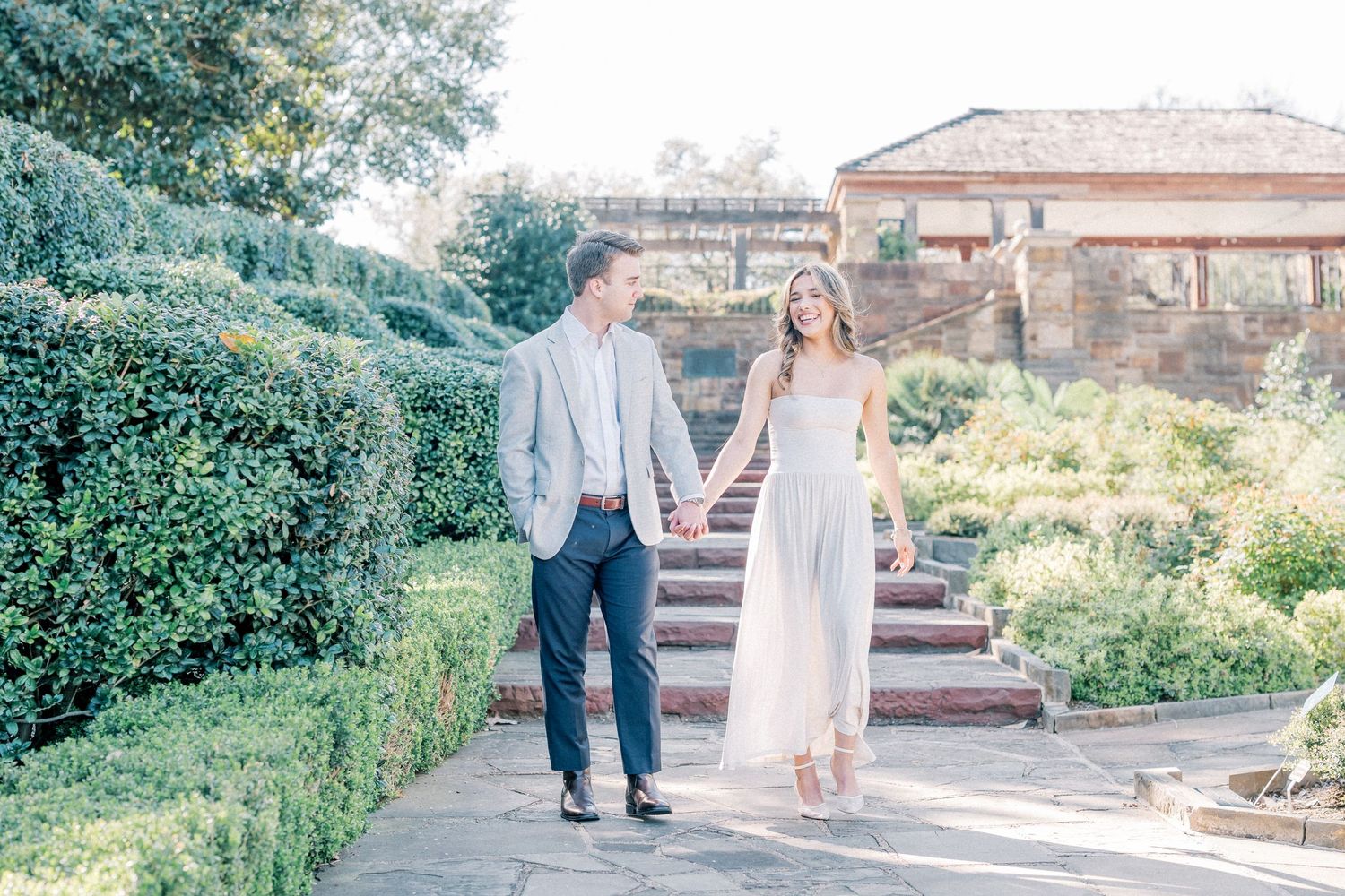A couple walks hand in hand along a garden path surrounded by lush greenery and stone steps on a sunny afternoon.