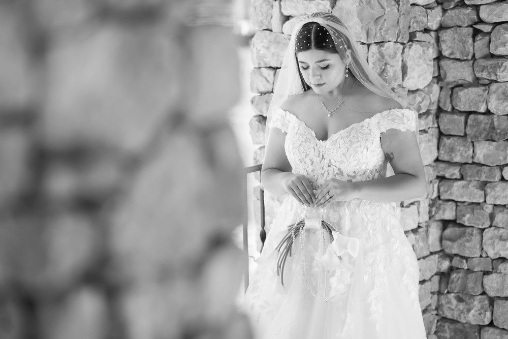 A bride in an elegant white gown stands thoughtfully in a stone colonnade.