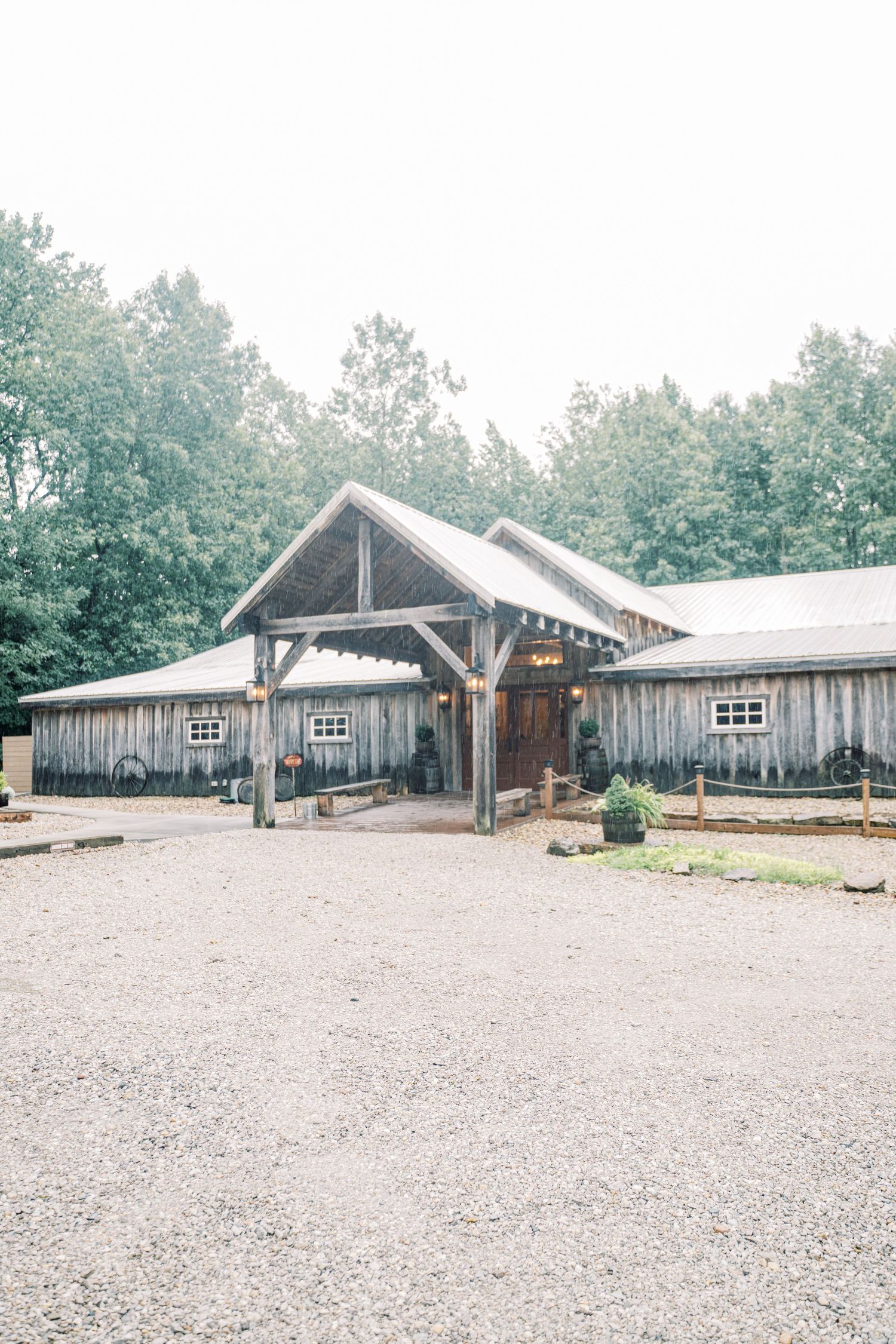 Rustic wooden barn wedding venue with gravel entrance, covered porch, and lush green trees in background.