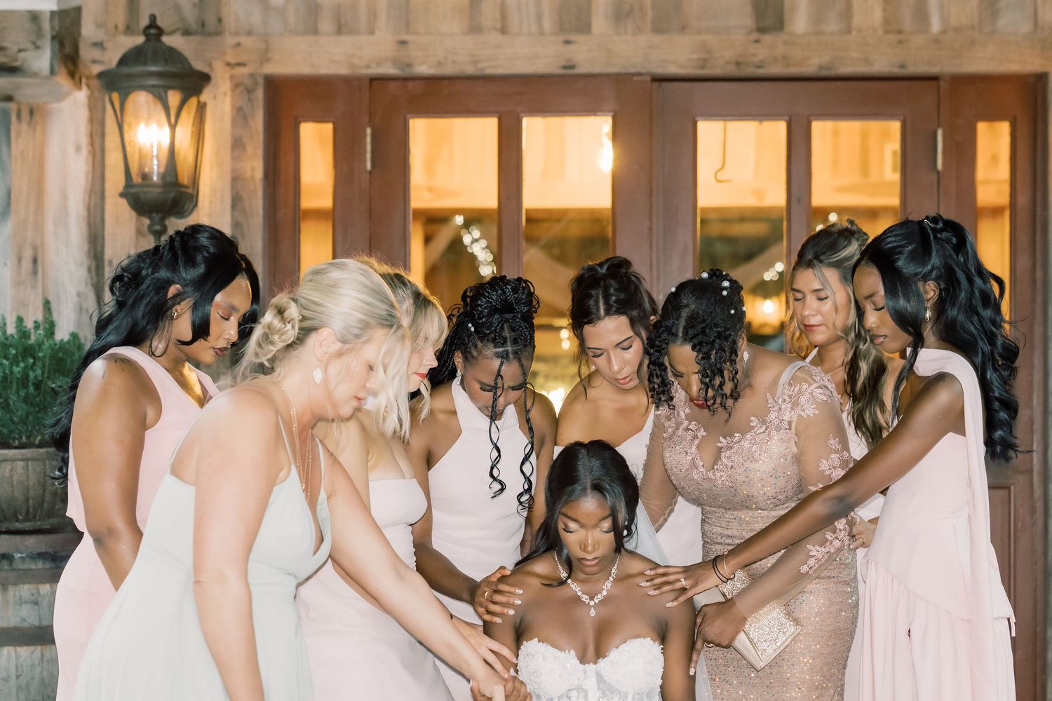 Bridesmaids in blush and white dresses gather around bride in prayer circle at elegant wedding venue.