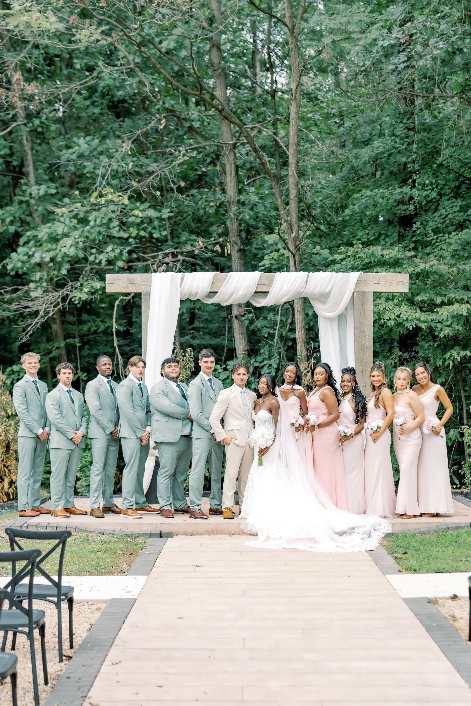 Wedding party poses under white draped arch, groomsmen in sage suits and bridesmaids in blush pink gowns outdoors.