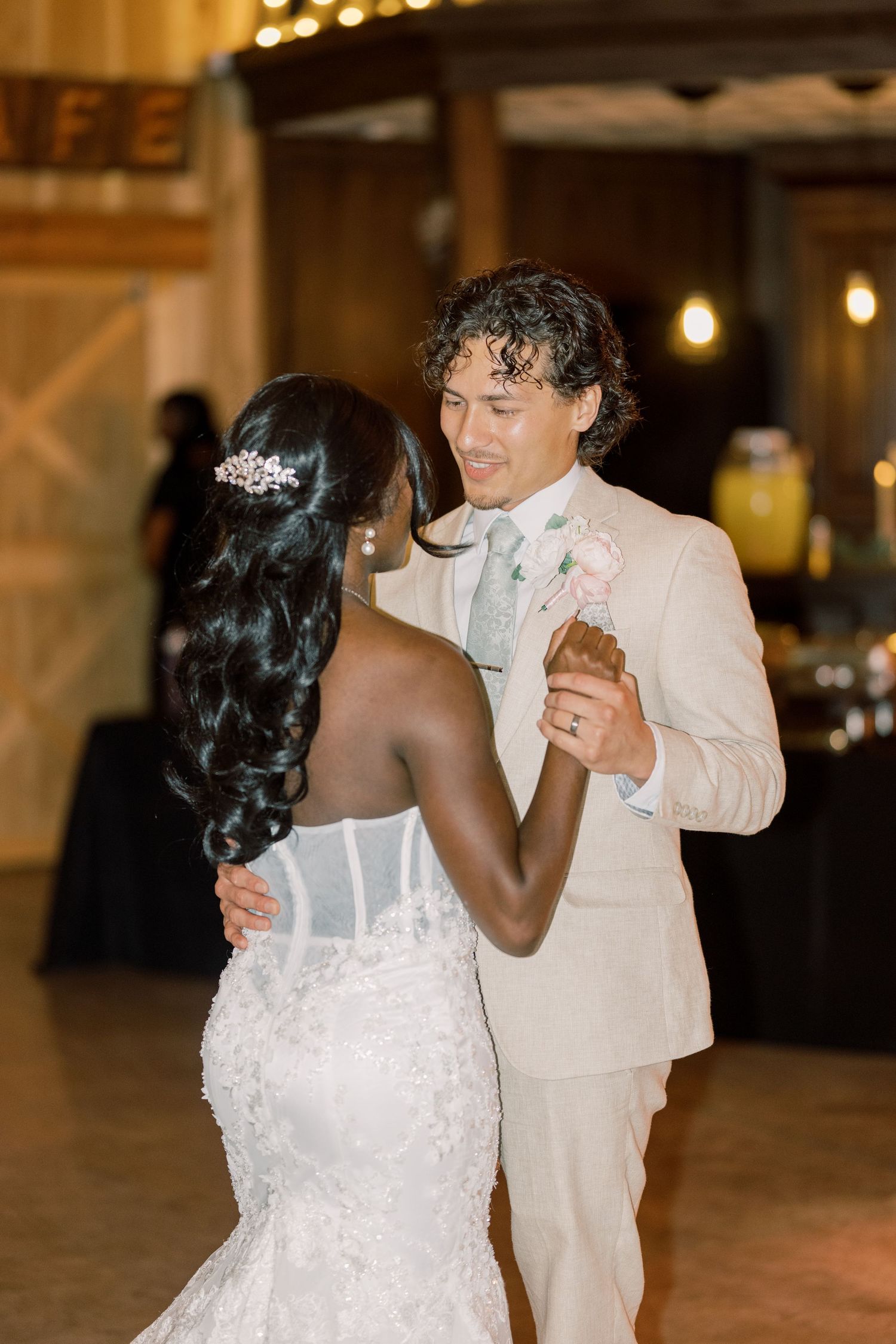 Interracial couple shares first dance, bride in lace mermaid gown and groom in tan suit.