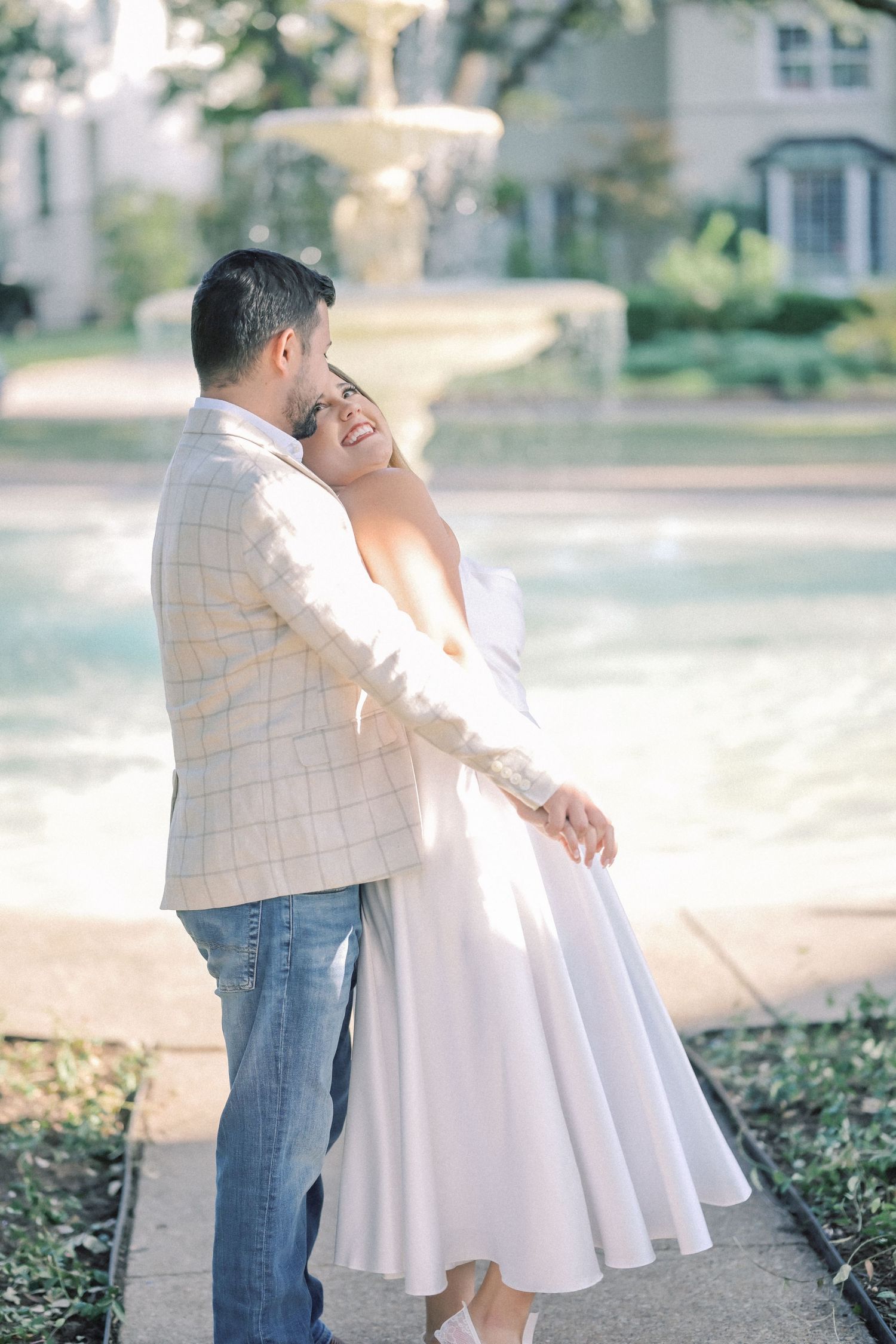 Couple in an affectionate embrace beside a sunlit park fountain, the man in a plaid blazer and jeans, the woman in a flowing white dress.