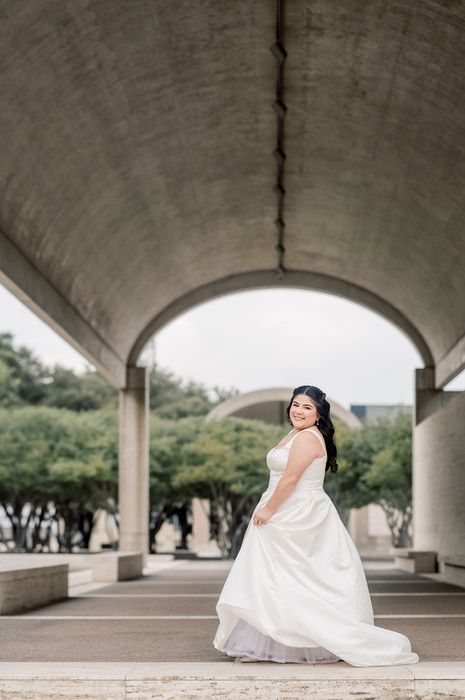 Smiling bride in elegant white gown posing under concrete arched walkway during outdoor bridal portraits.