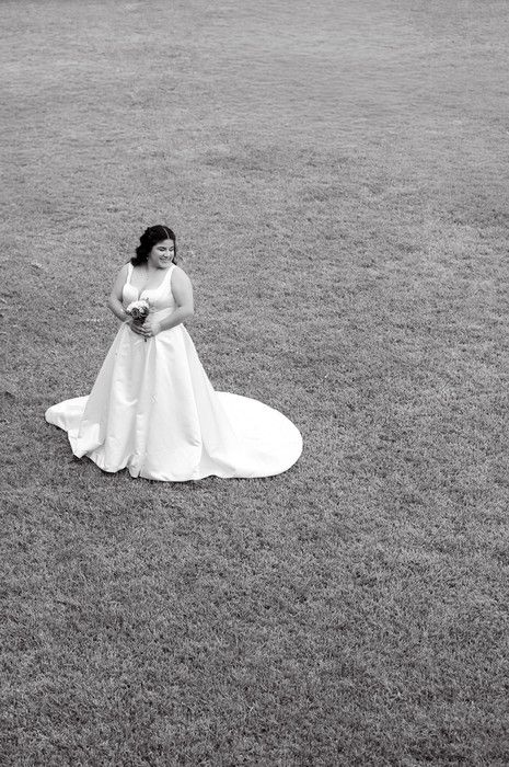 Black and white photo of a bride in a flowing white gown with train, holding bouquet on a grass lawn.