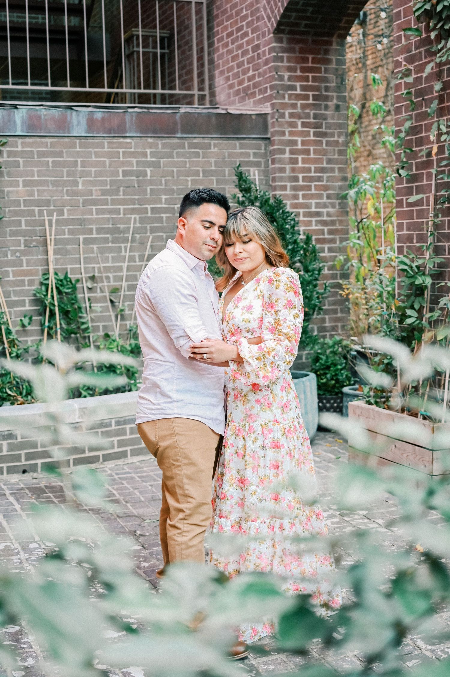 Couple embracing tenderly in a lush garden courtyard, woman in floral dress, man in casual attire.