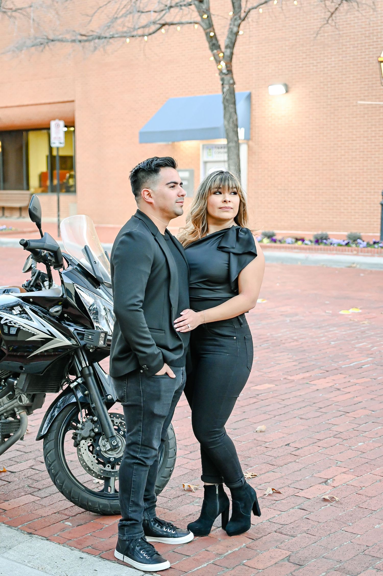 Couple in matching black outfits posing by a motorcycle on a brick sidewalk.