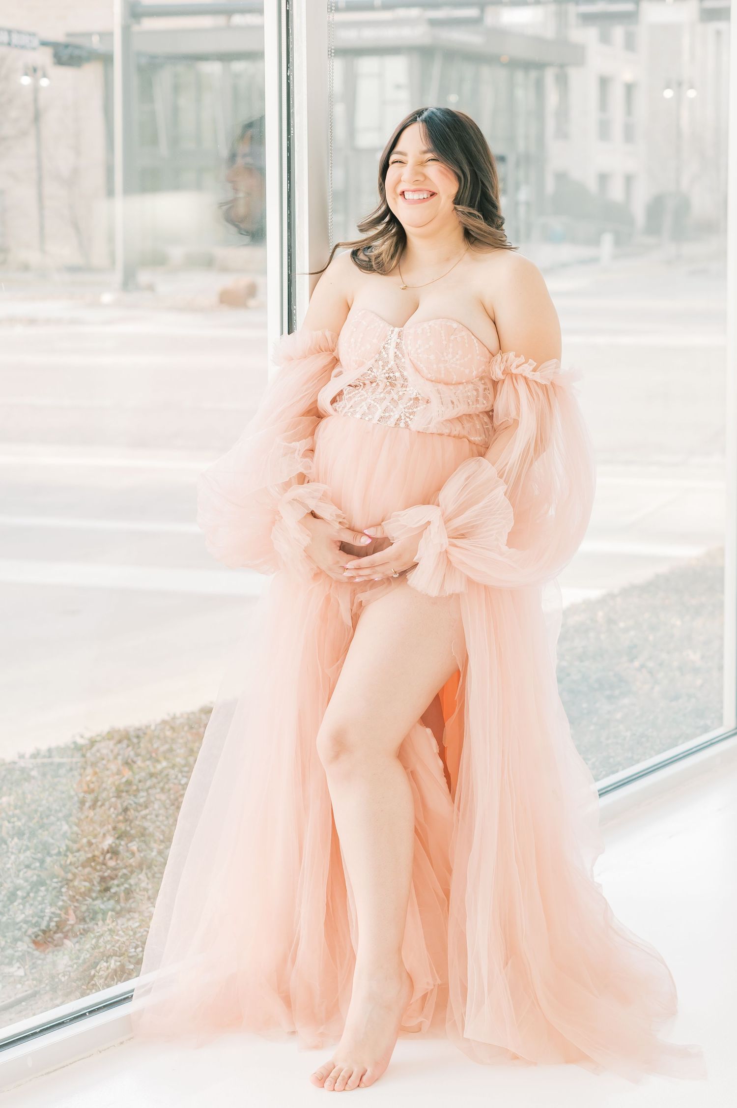 Smiling pregnant woman in a blush pink tulle maternity gown standing by a large window, cradling her baby bump.