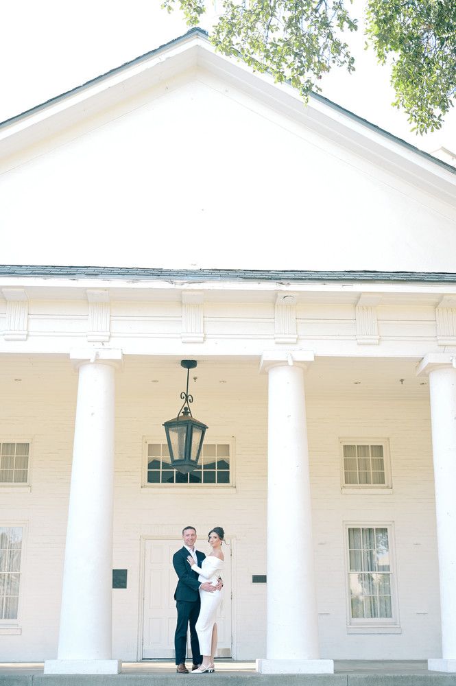 Bride and groom posing before a white colonial building with tall columns and a hanging lantern on their wedding day.