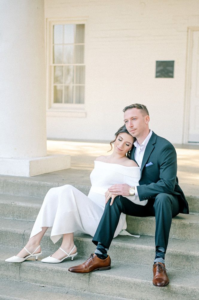 Bride in white off-shoulder gown sits between groom's legs on stone steps of a classic white building.