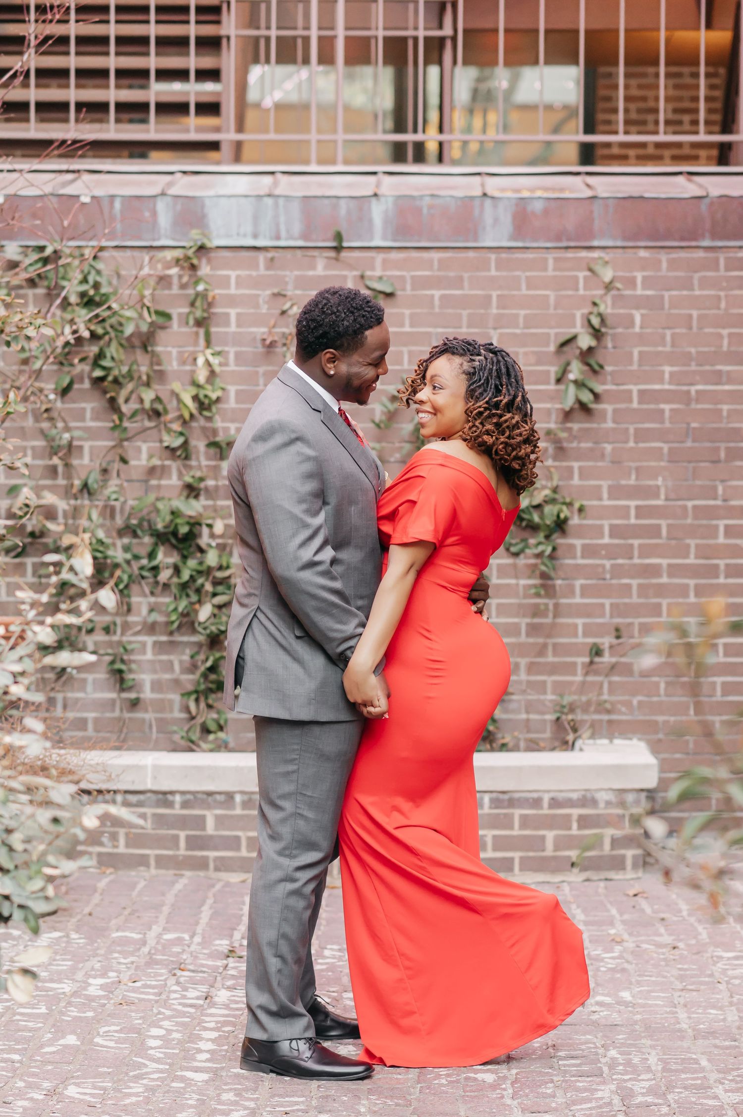 Couple in love posing together, man in gray suit and woman in elegant red gown, against a brick wall backdrop.