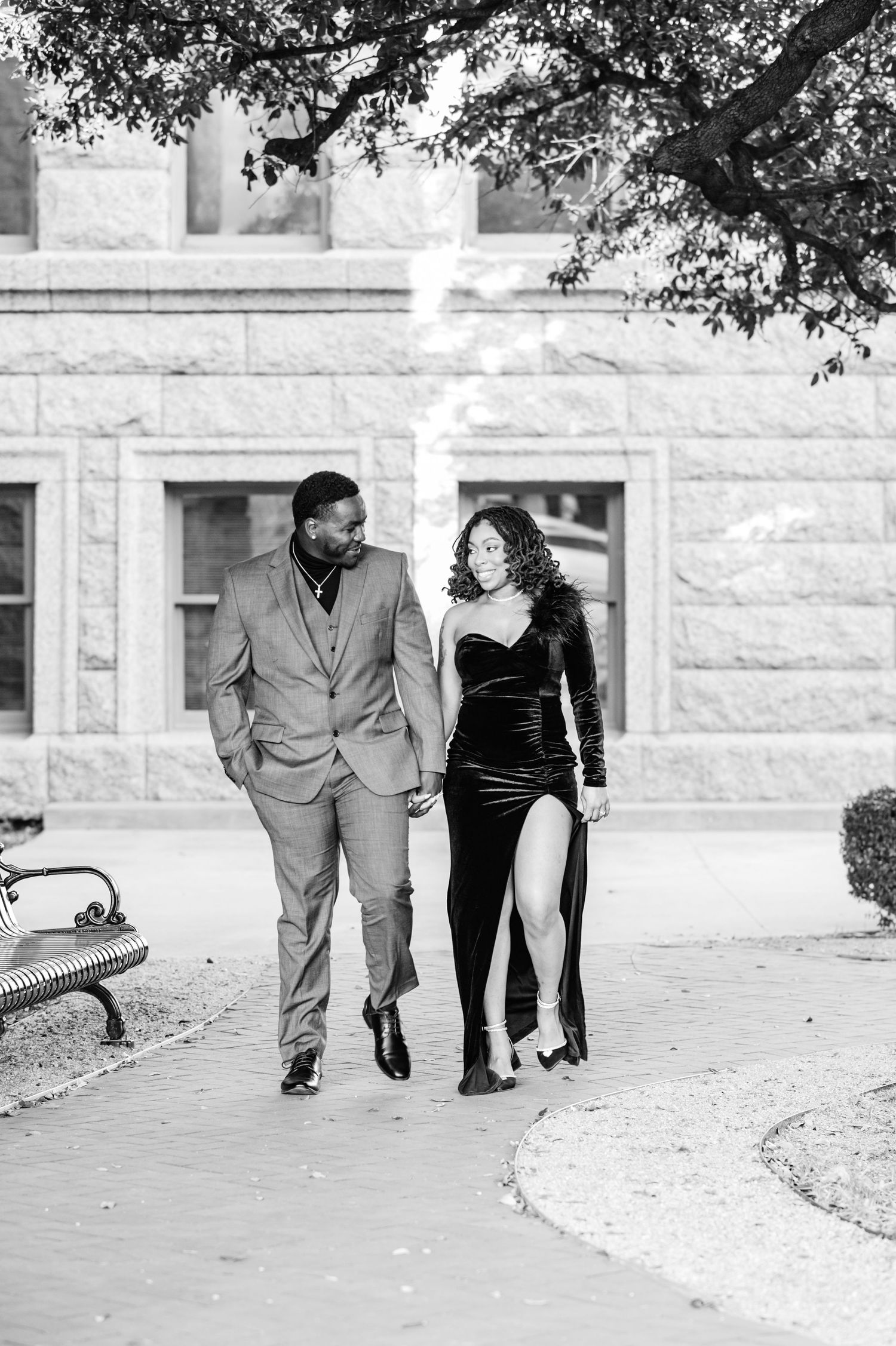 Couple walking hand-in-hand in black and white, man in suit and woman in black velvet dress, near a stone building.