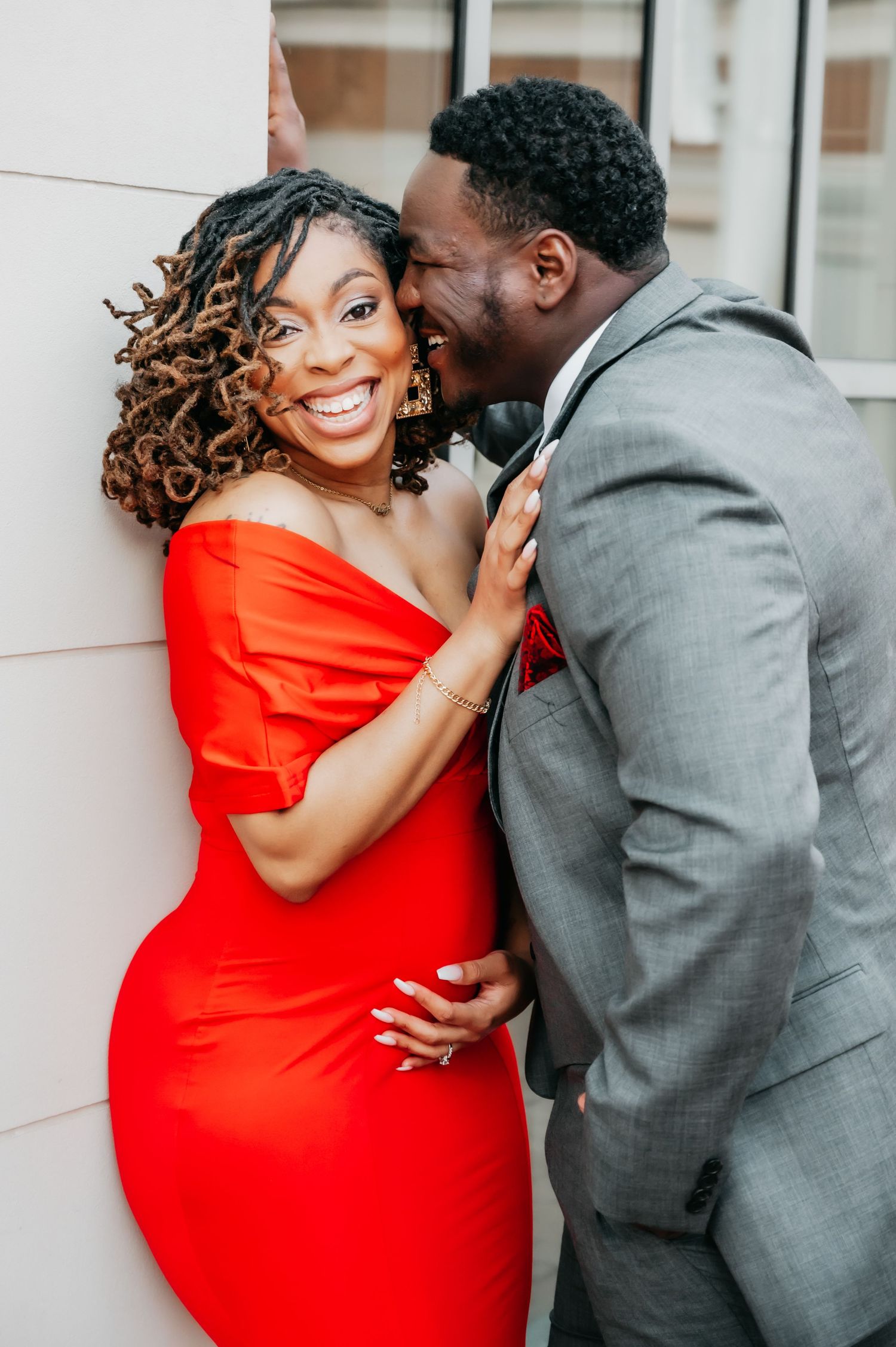 A joyful couple poses together, woman in red off-shoulder dress laughing as man in gray suit whispers in her ear.