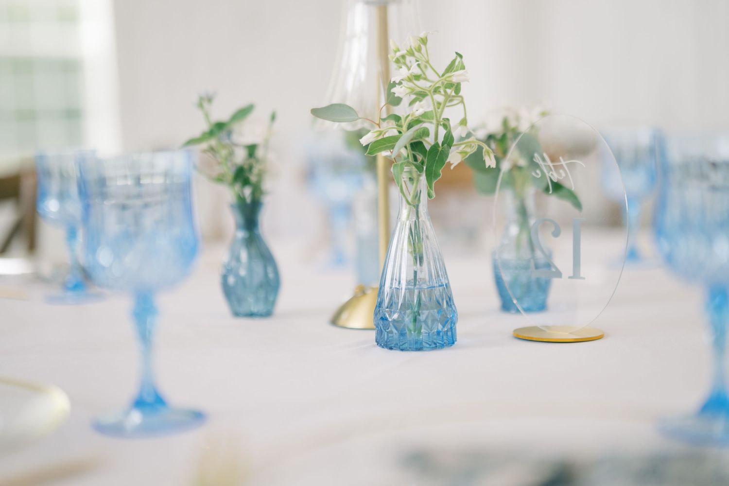 Elegant wedding reception table with blue glassware and small bud vases with greenery on a white tablecloth.