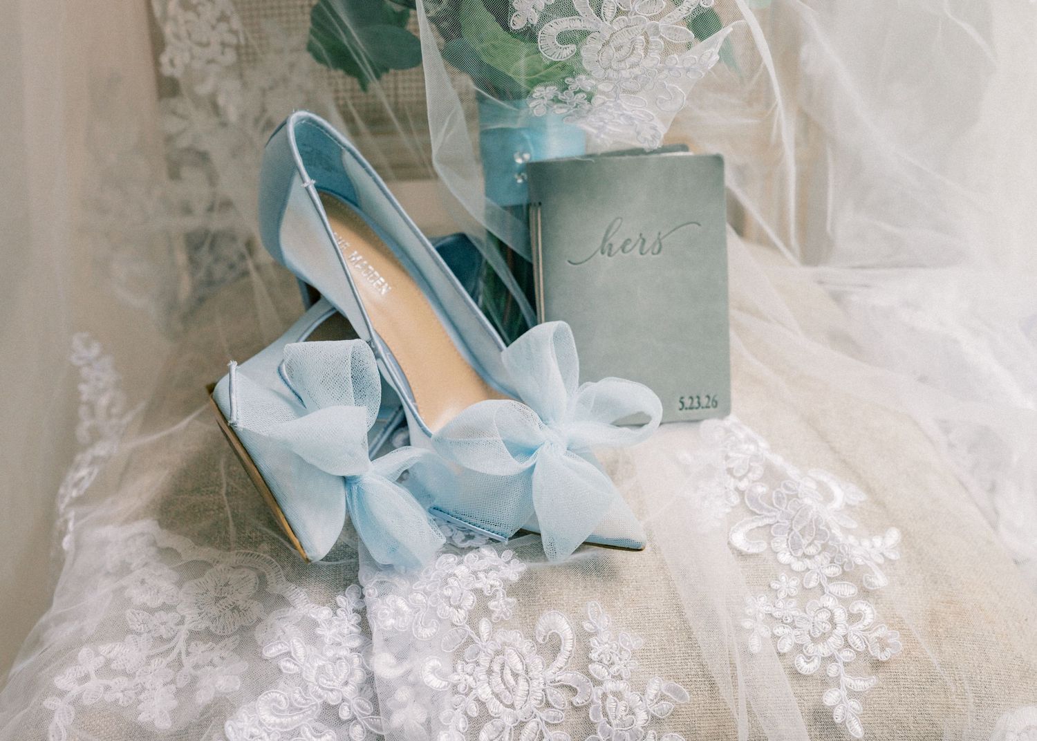 Light blue bridal heels with bow detail beside a vow book on white lace wedding dress.