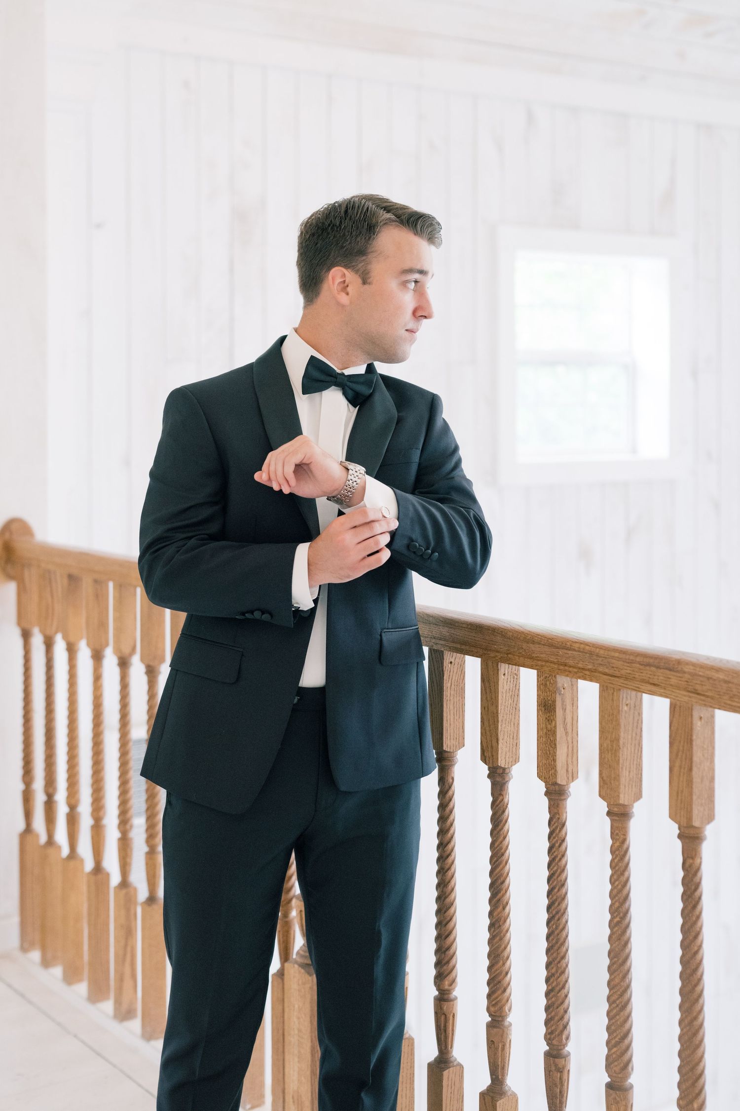 Groom in black tuxedo with bow tie adjusts cufflinks near wooden staircase railing in white interior.
