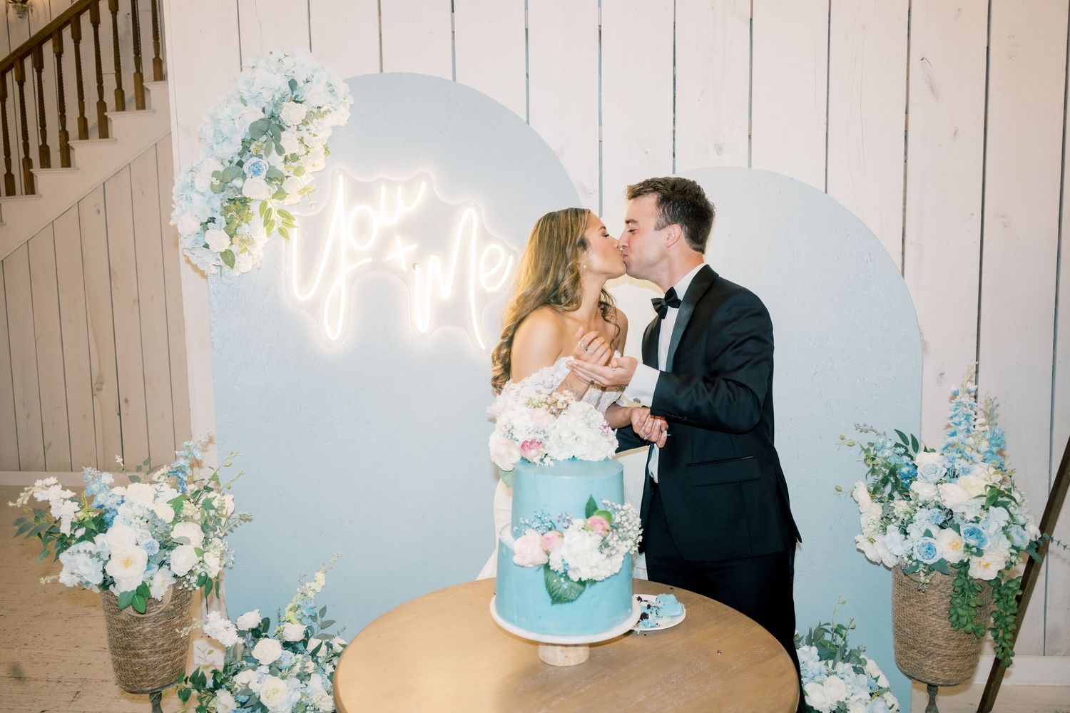 Newlyweds kissing behind a blue wedding cake in front of a pastel arch with neon sign.