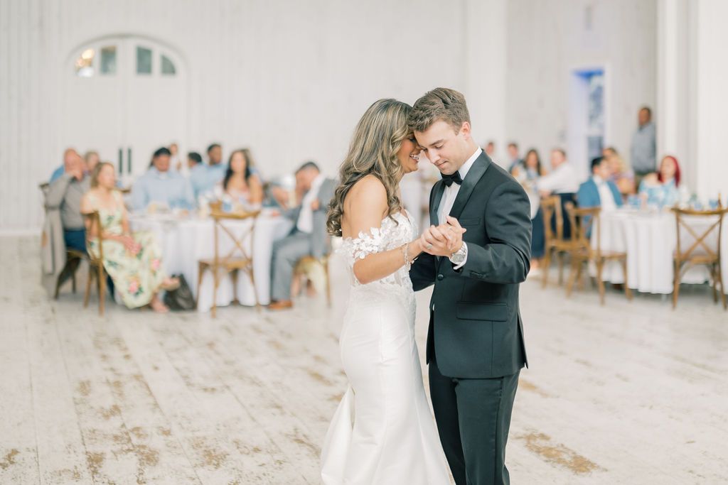 Bride and groom share their first dance in an elegant white reception hall with guests seated at tables in the background.