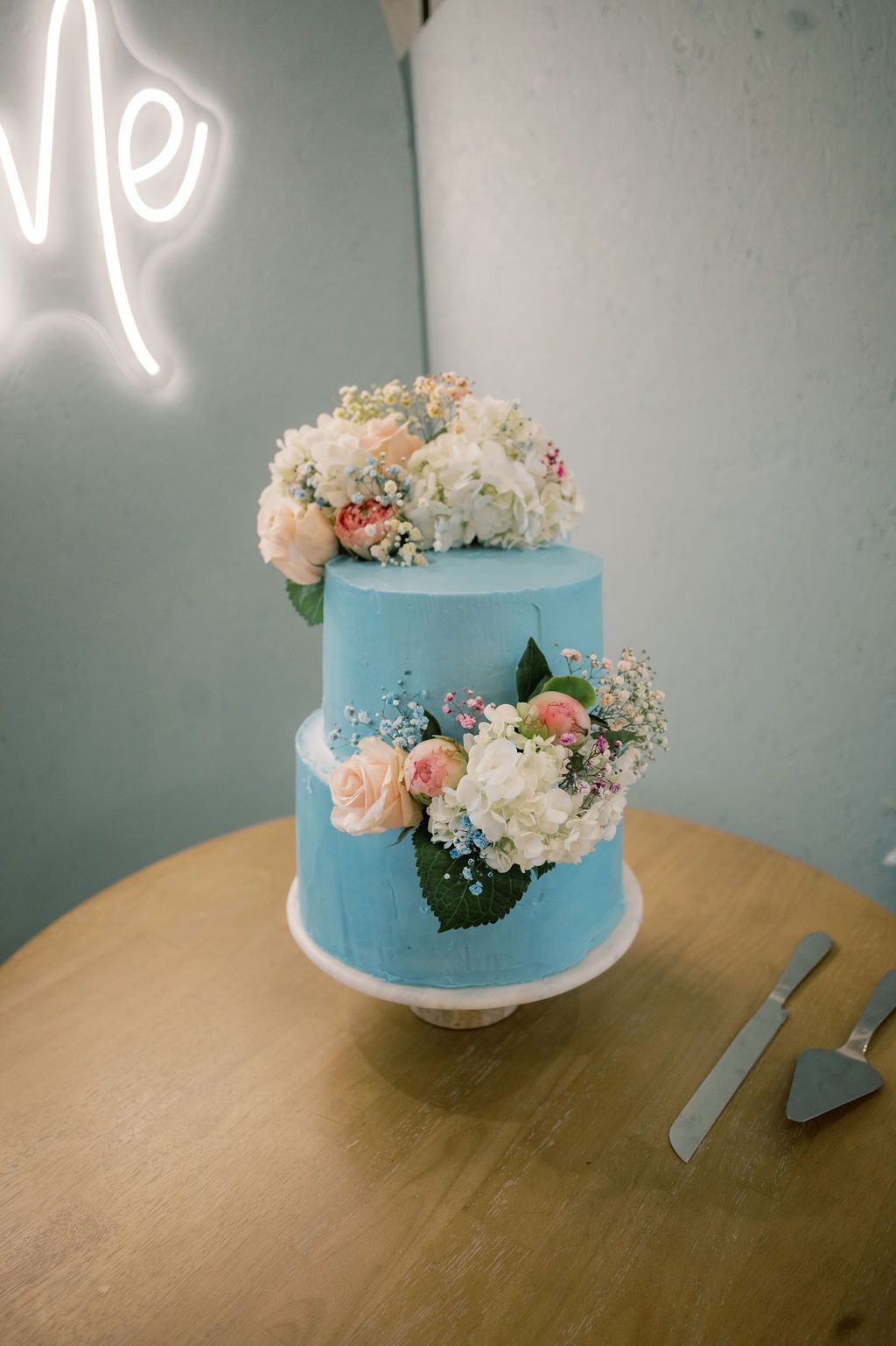 Two-tier blue wedding cake decorated with fresh pink roses and white hydrangeas on a wooden table.