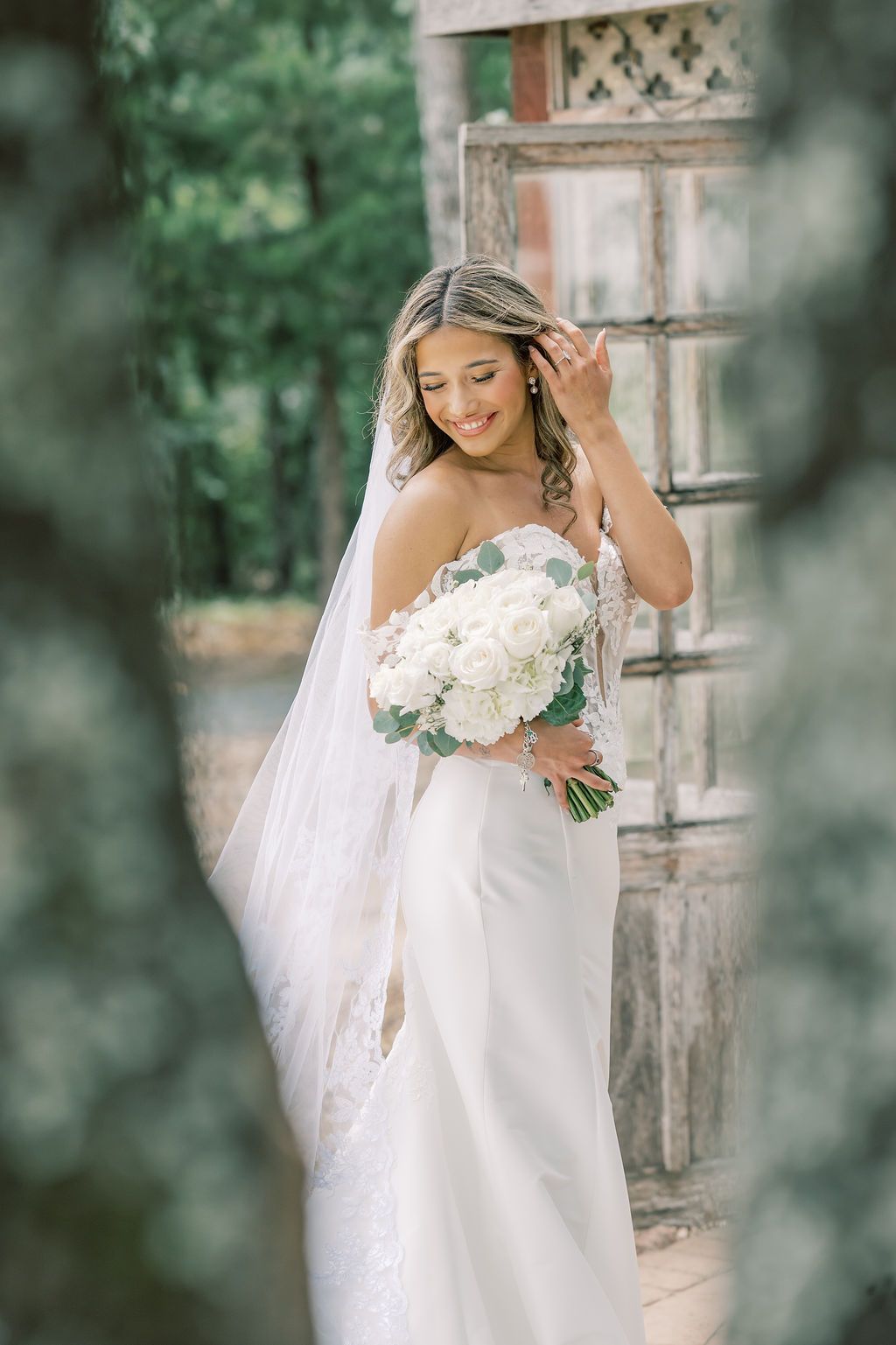Smiling bride in white off-shoulder gown holds white hydrangea bouquet, wearing veil outdoors near rustic wooden door.