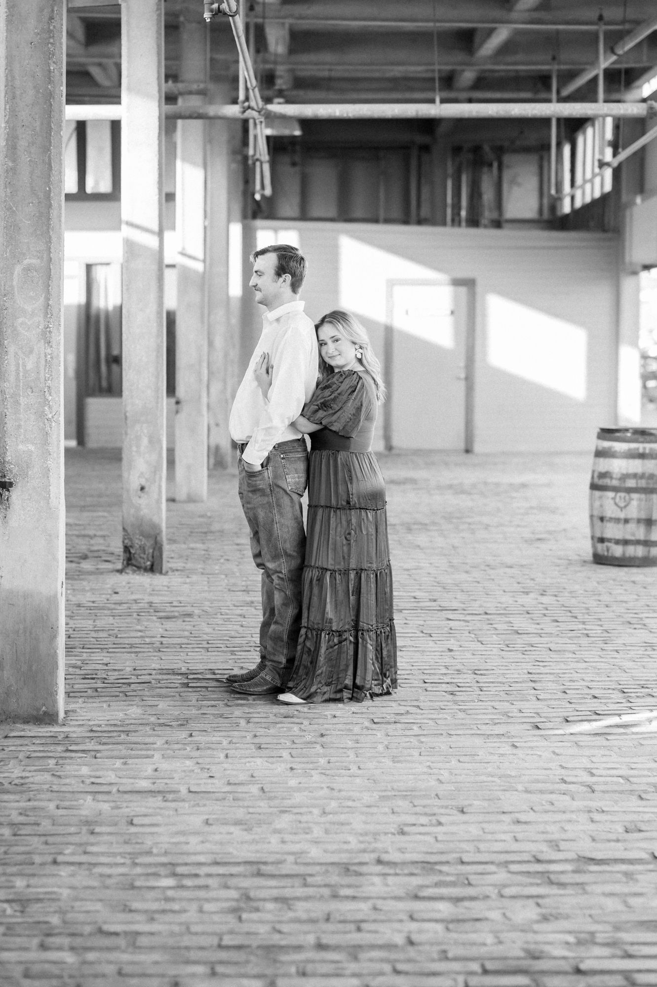 Black and white portrait of a couple embracing in an industrial space with brick floors and exposed beams.