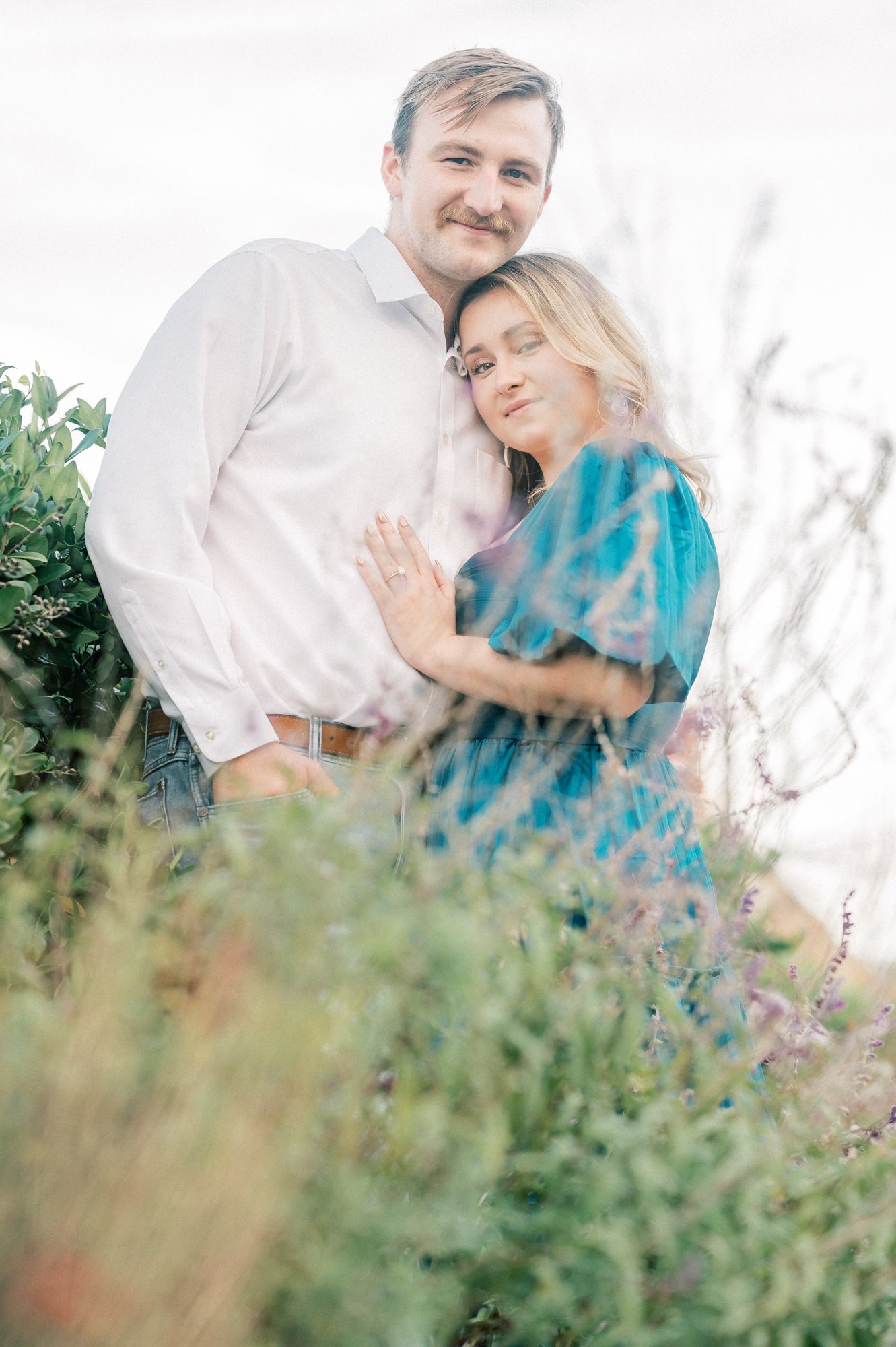 A romantic couple embraces in an outdoor setting with soft natural lighting and foliage in the background.