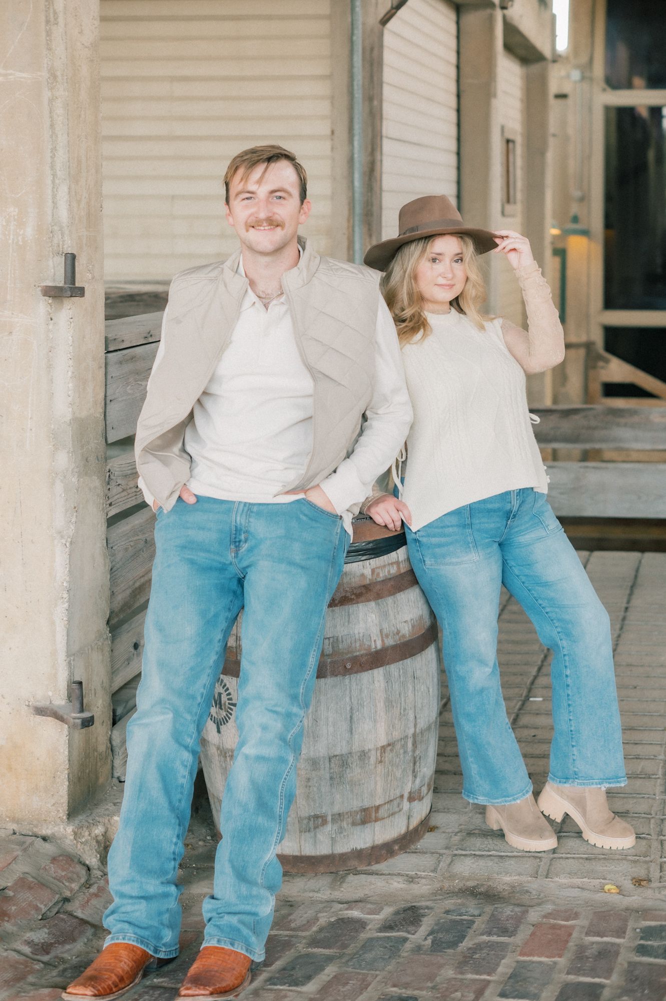 Couple sitting together on a rustic wooden barrel against a white building exterior.