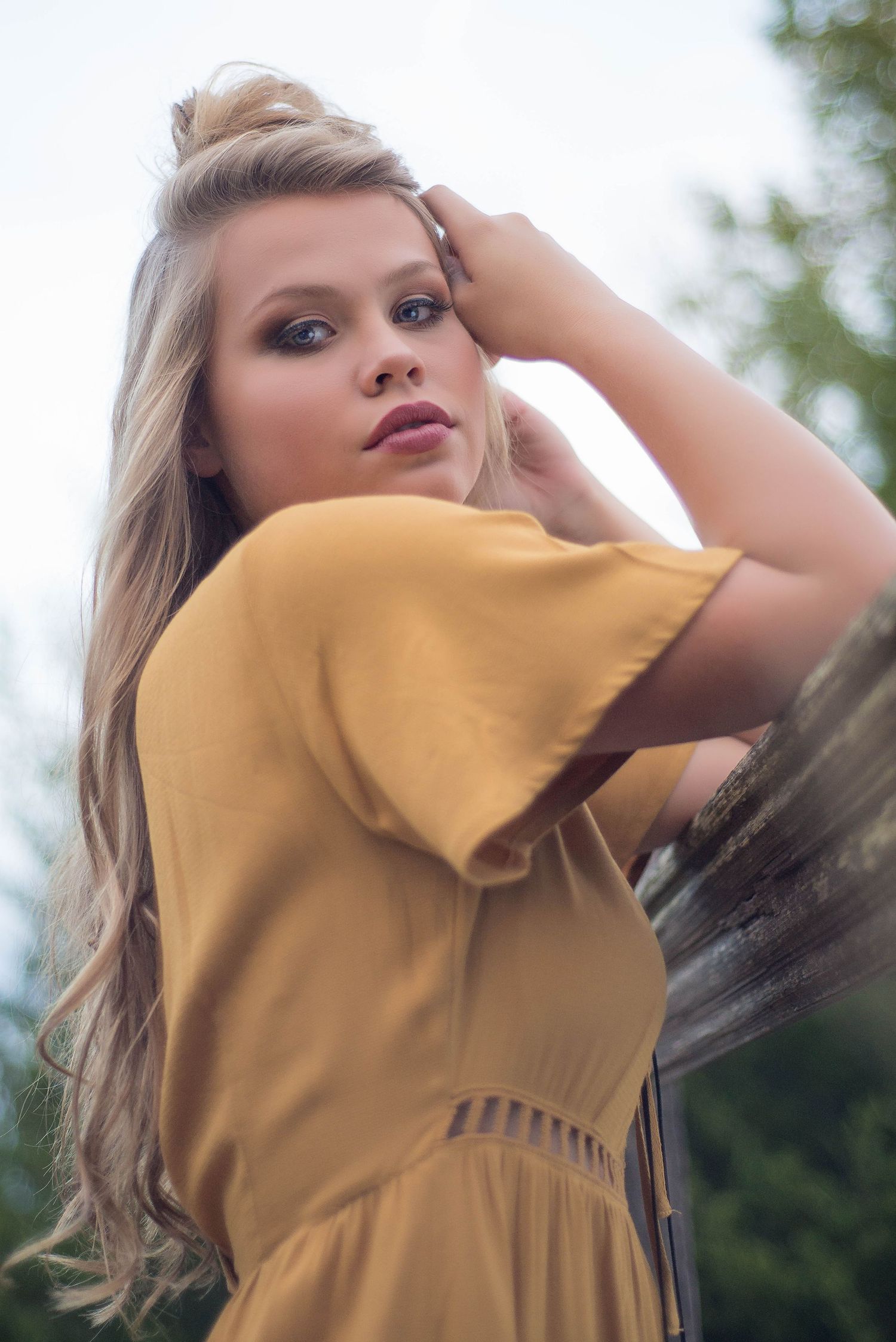 Portrait of someone in a mustard yellow dress with soft makeup and flowing blonde hair against a blurred outdoor background.
