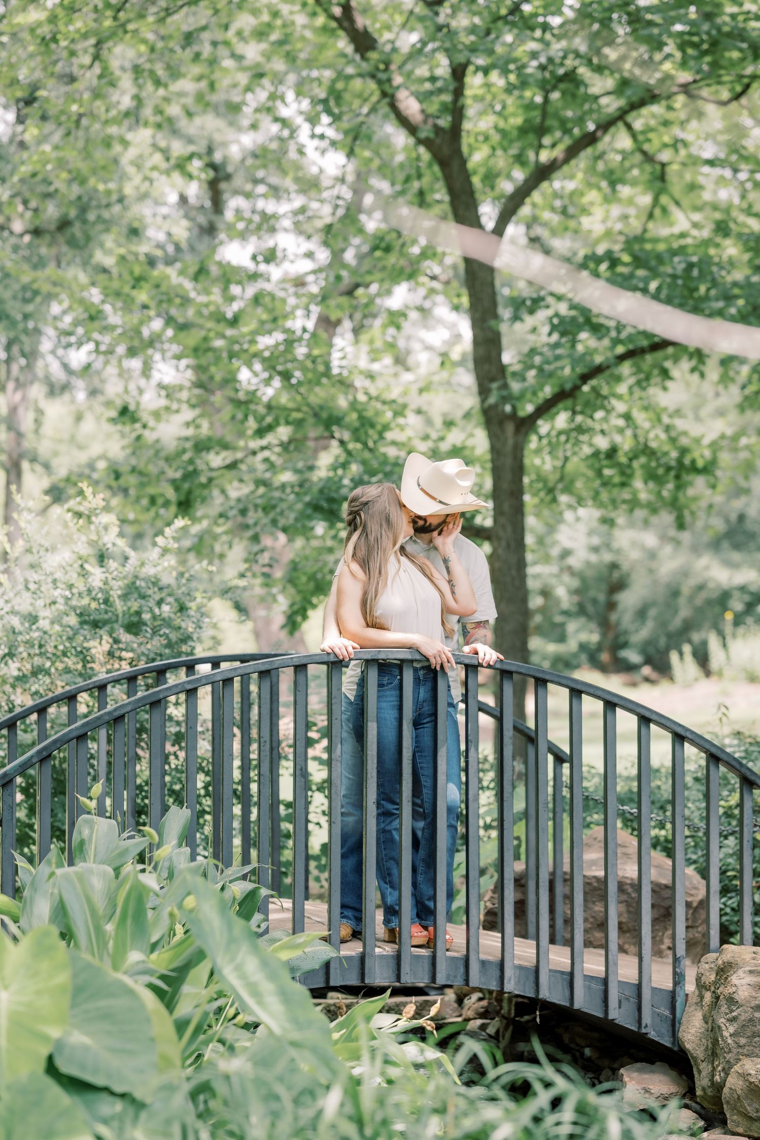 Romantic couple kissing on an arched bridge surrounded by lush green trees and garden foliage.