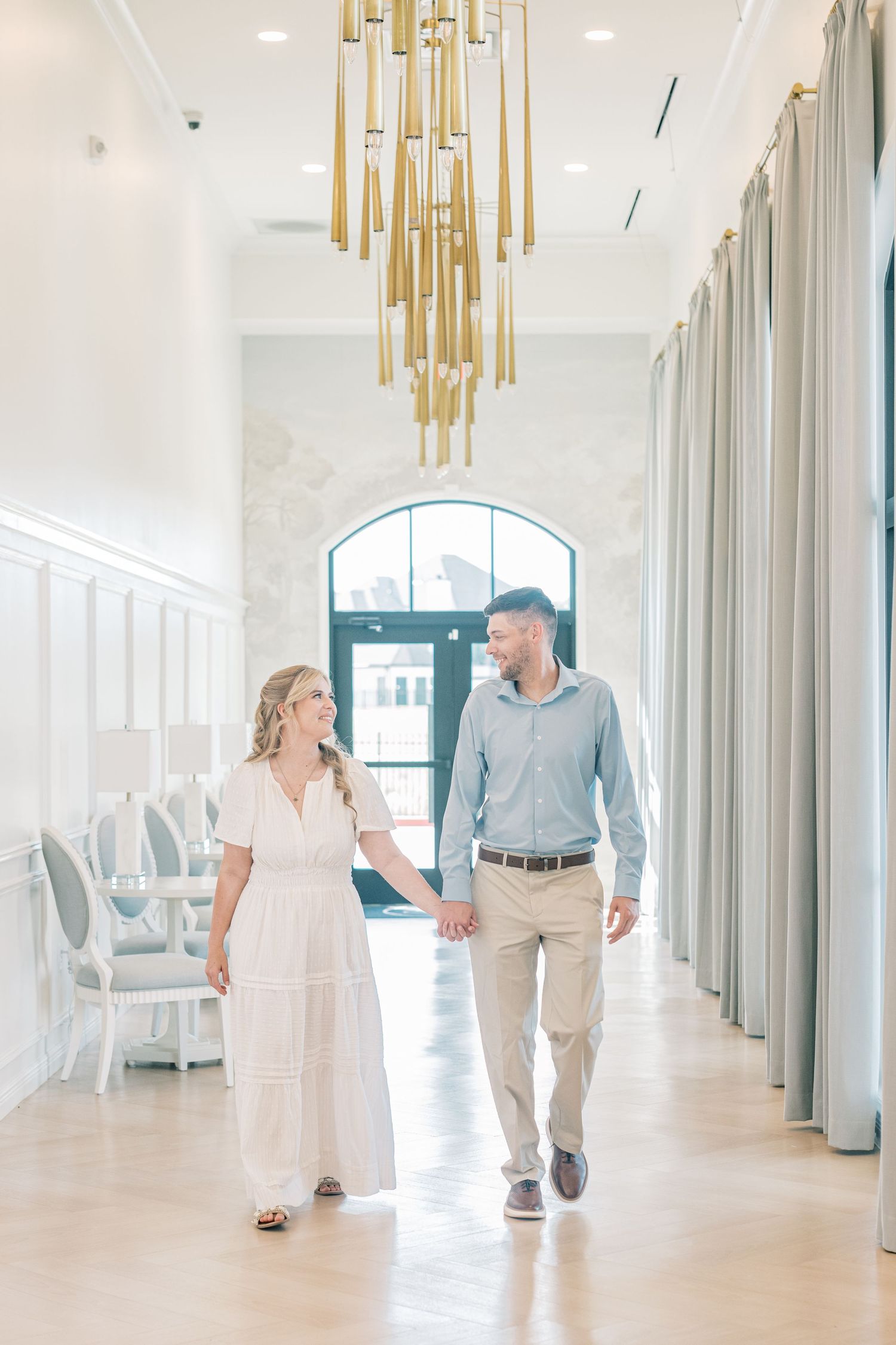 Couple holding hands walking through elegant white hallway with gold chandelier during engagement session.