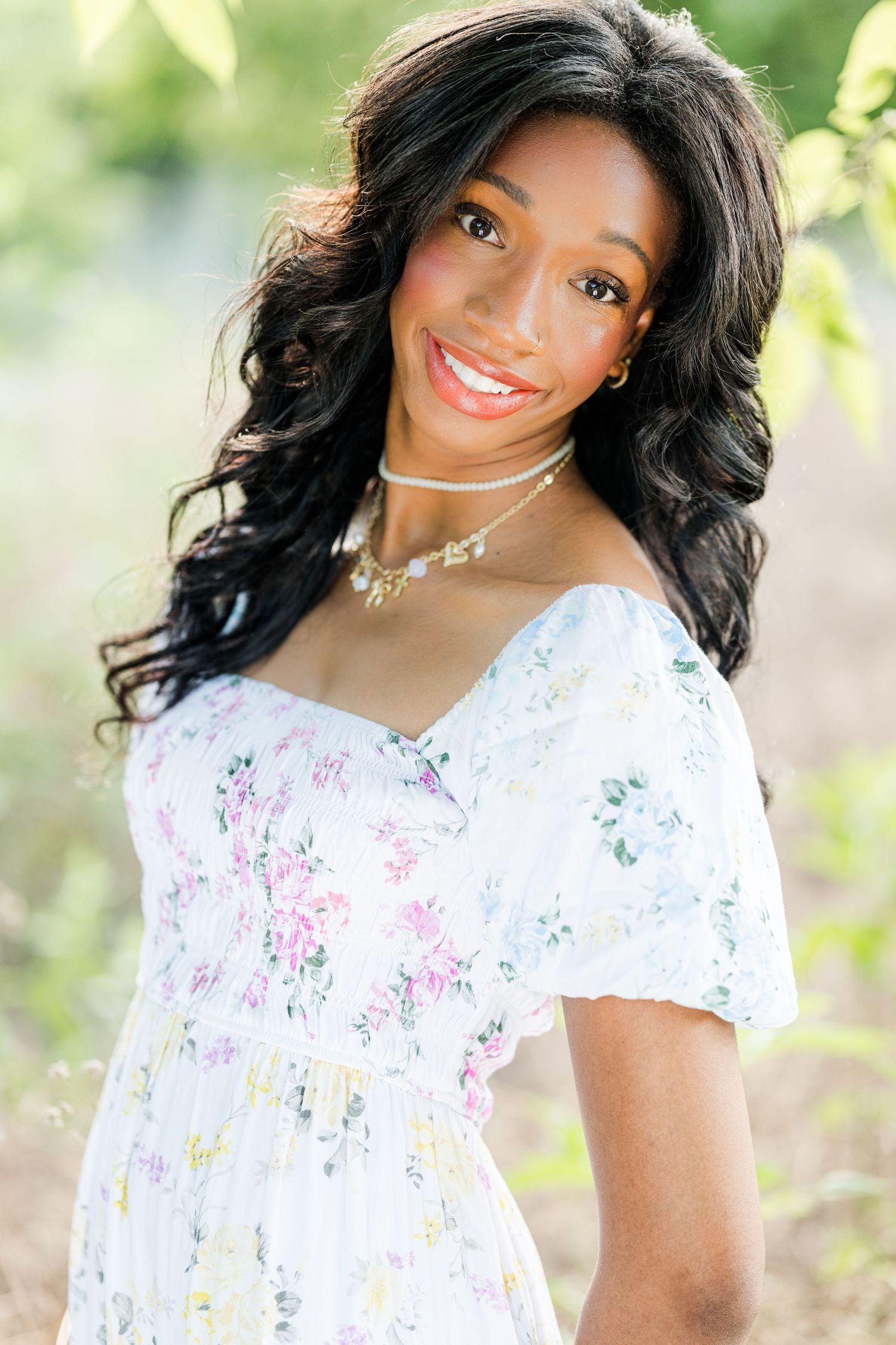 Smiling young woman in white floral off-shoulder dress with layered necklaces, posing outdoors among green foliage.