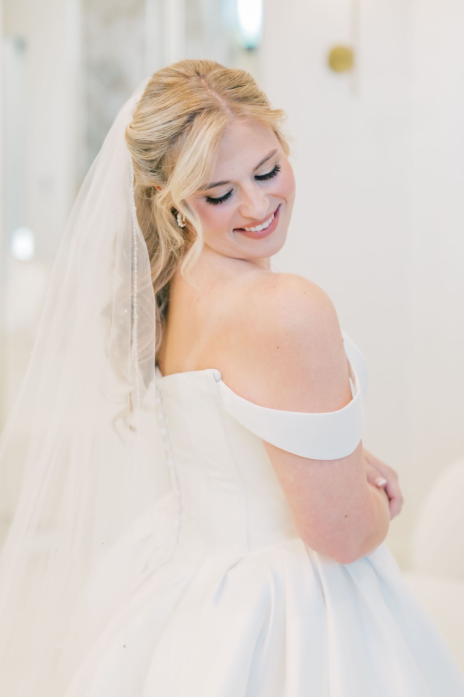 Smiling blonde bride in an off-shoulder white gown and veil looking down in a bright bridal suite.