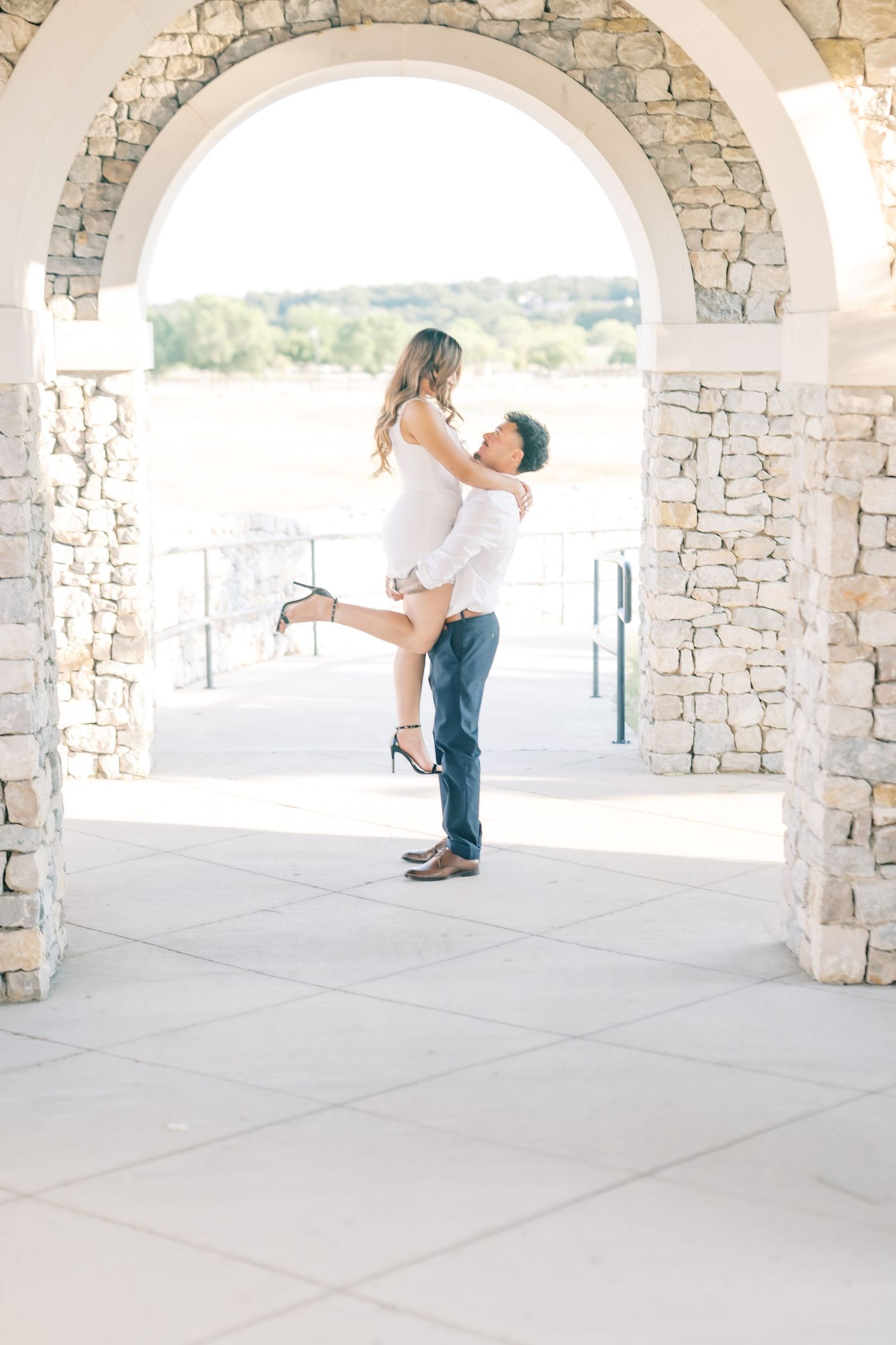 Man lifting woman in white dress under a stone archway during an outdoor engagement photo session.