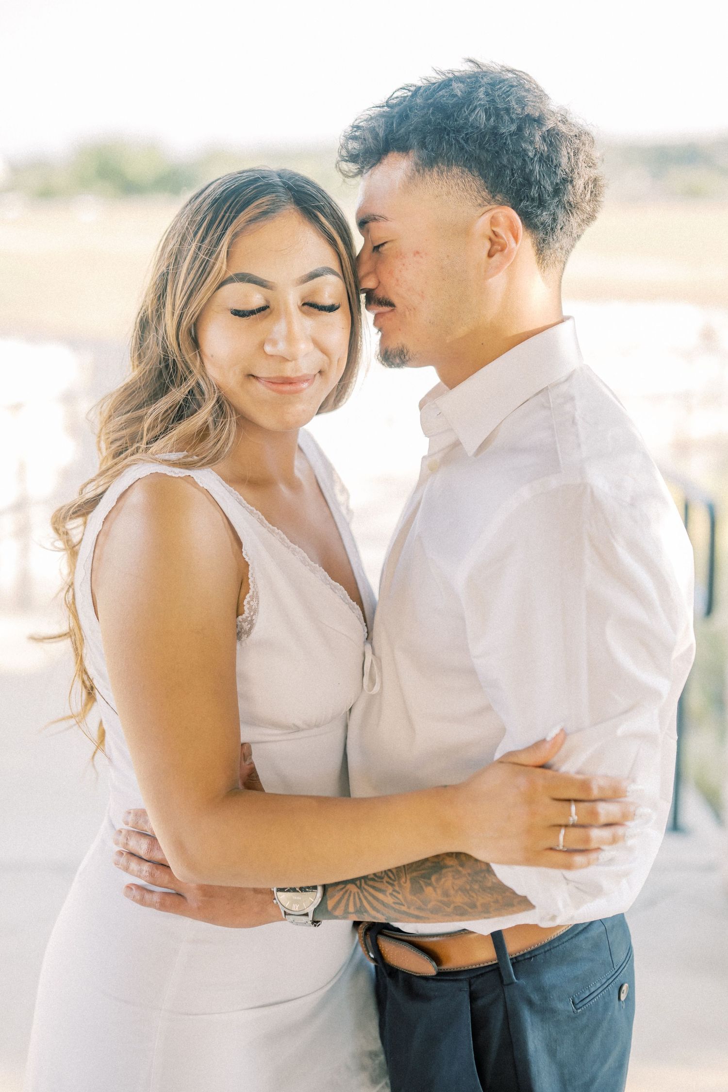 Couple embracing tenderly outdoors, woman in white dress smiling softly as man nuzzles her cheek during engagement session.