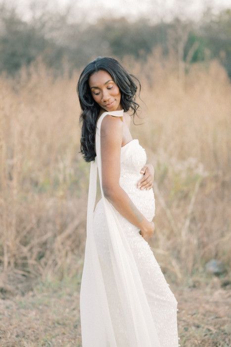 Pregnant woman in white dress cradles her baby bump while standing in a sunlit field.