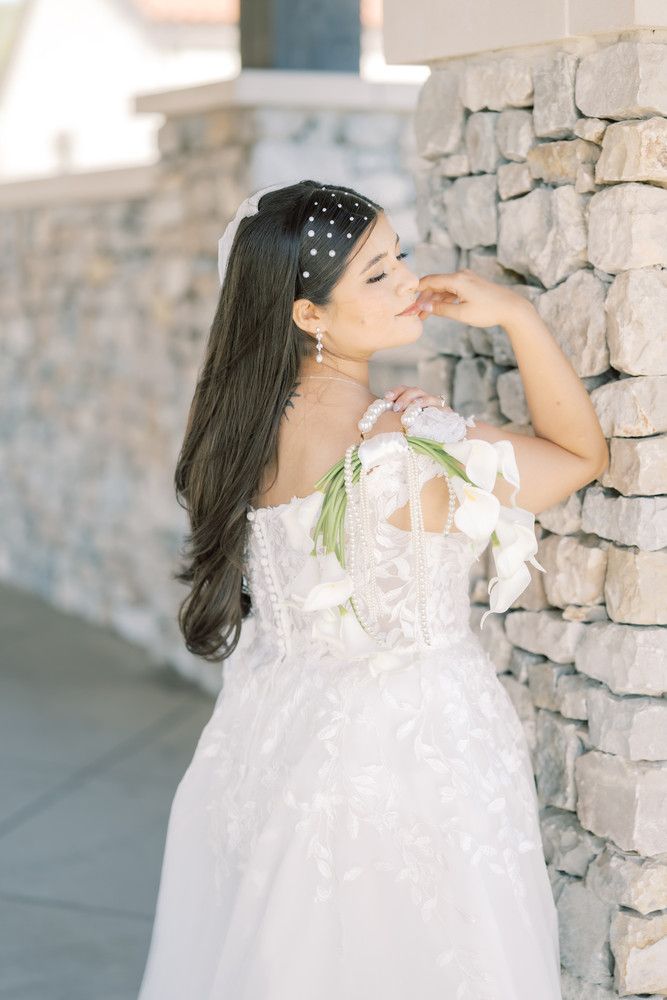 A bride poses gracefully against a stone wall in a flowing white gown with floral accents.