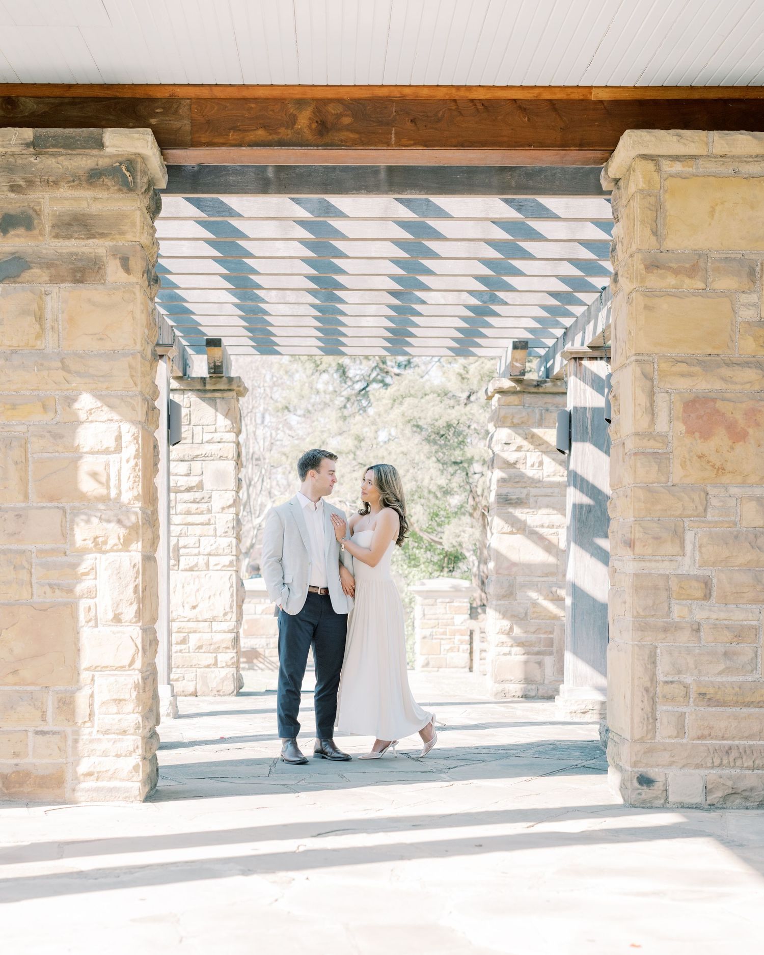 A couple shares a romantic moment under a covered walkway with stone pillars and natural lighting.