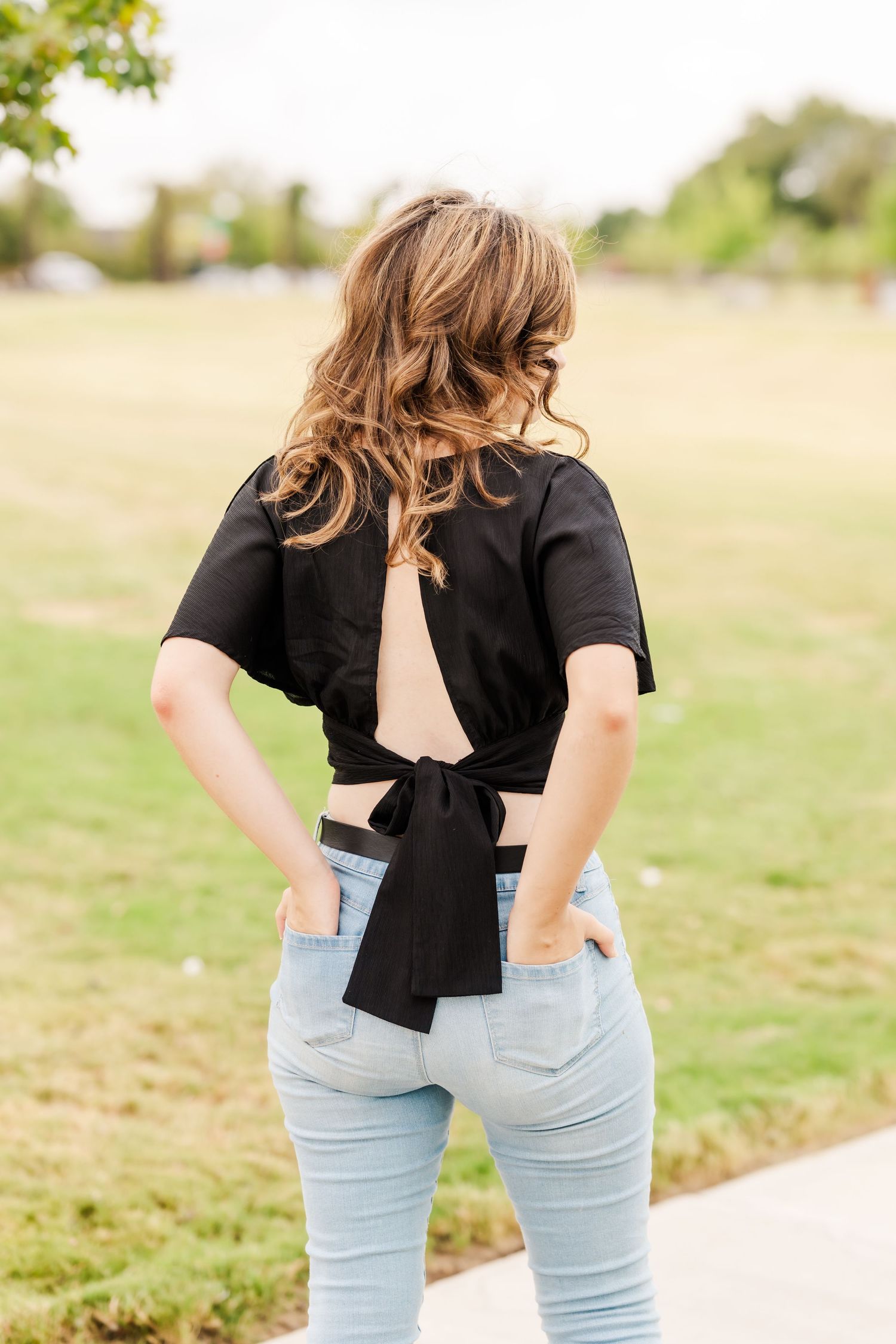 Woman in light blue jeans and black open-back tie crop top standing outdoors in a park.