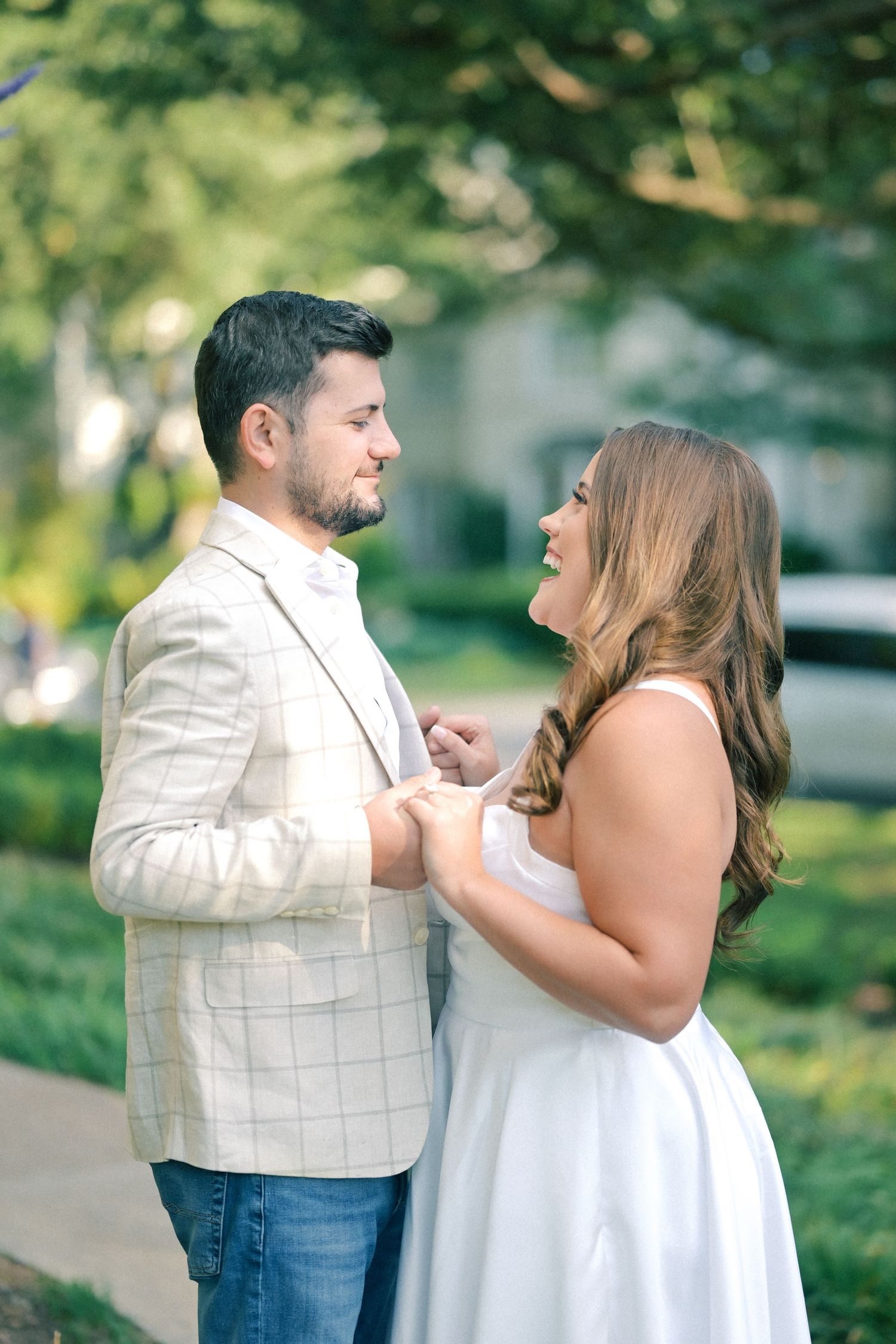 A smiling couple holds hands and gazes at each other during an outdoor engagement photo session surrounded by green trees.